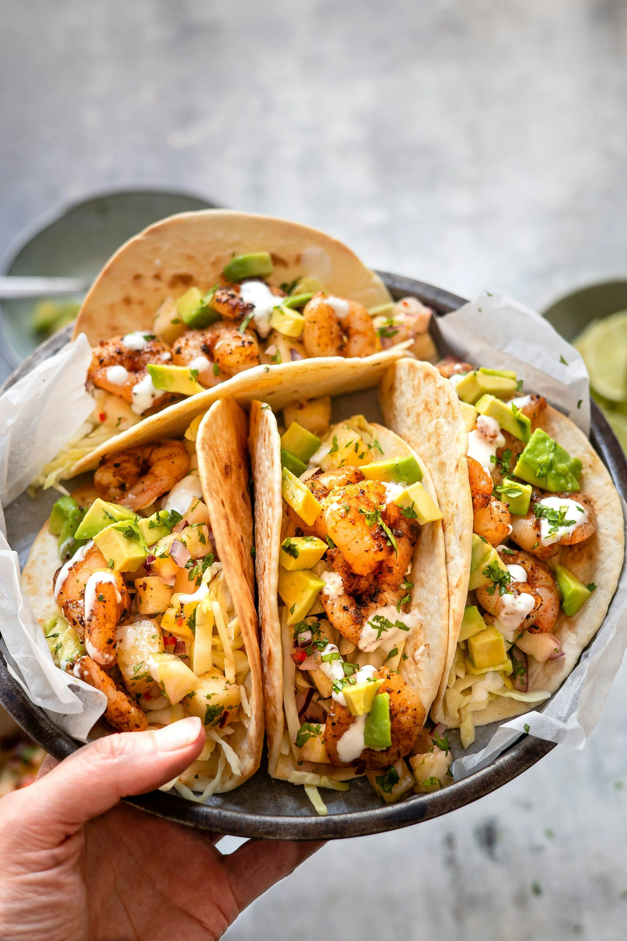 Three shrimp tacos topped with avocado, sauce, and herbs, served on a basket lined with paper.