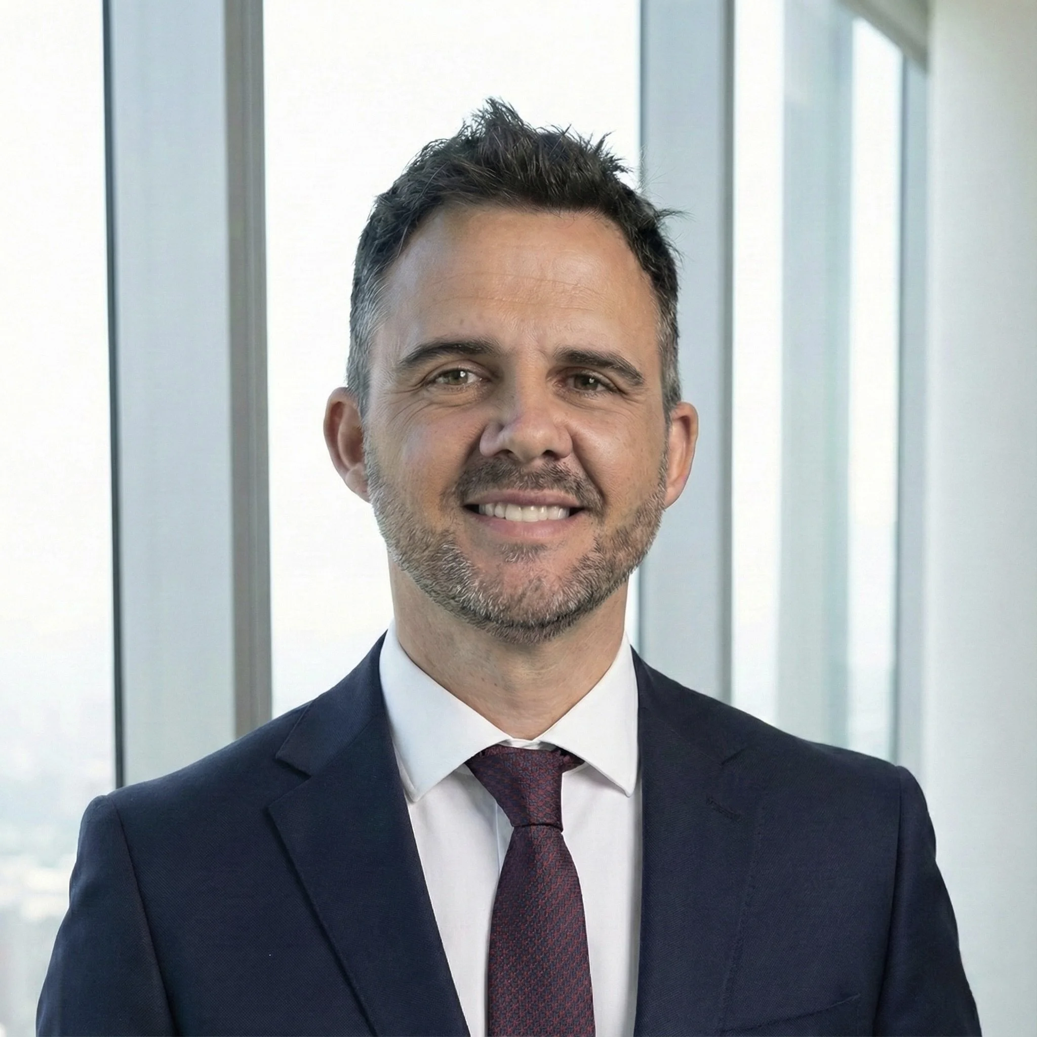 Hombre sonriendo, vestido con traje formal, en una oficina moderna con ventanas grandes y vista urbana.