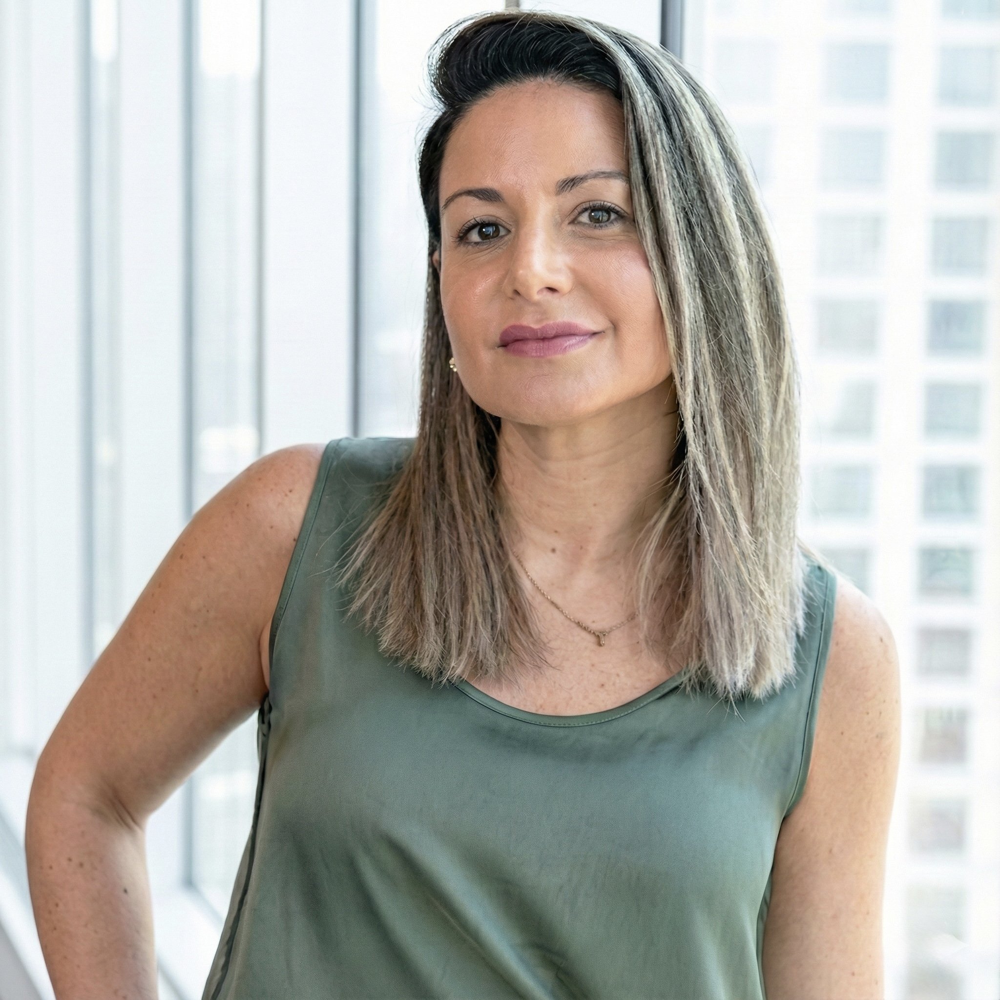 Retrato de una mujer con pelo castaño y rubio, usando una blusa verde y un collar, en un entorno con ventanas grandes y edificios altos al fondo.