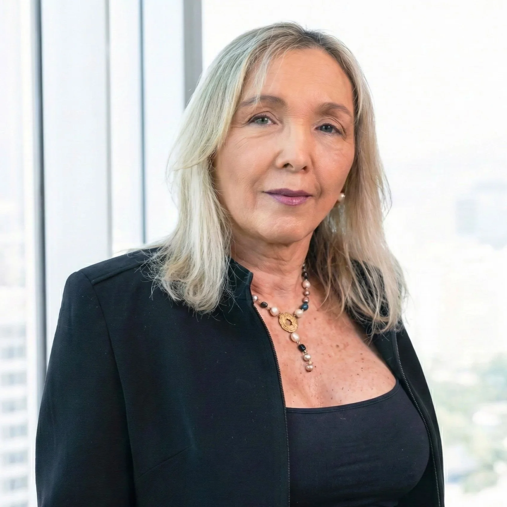 Mujer de cabello gris, vestido negro, con collar de perlas y fondo de ventanas