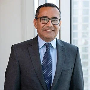 Retrato de un hombre joven con traje oscuro, camisa blanca y corbata azul, sonriendo en un entorno de oficina con ventanas y ciudad de fondo.