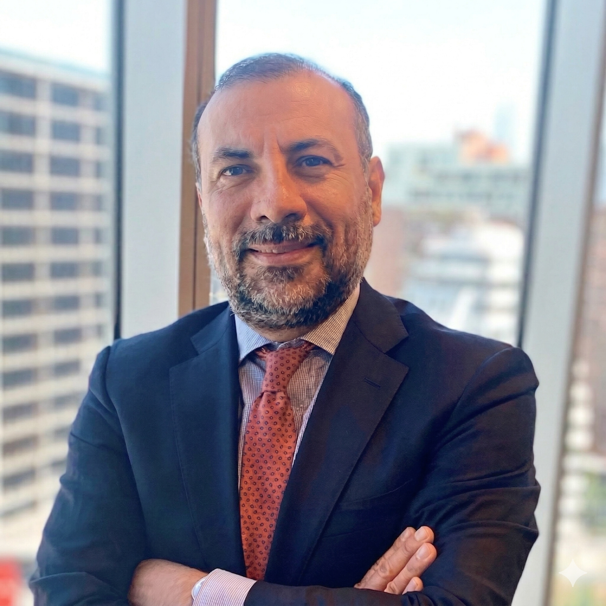 Retrato de un hombre con barba y cabello corto, vestido con traje oscuro y corbata anaranjada, en una oficina con ventanas grandes y vista de edificios.