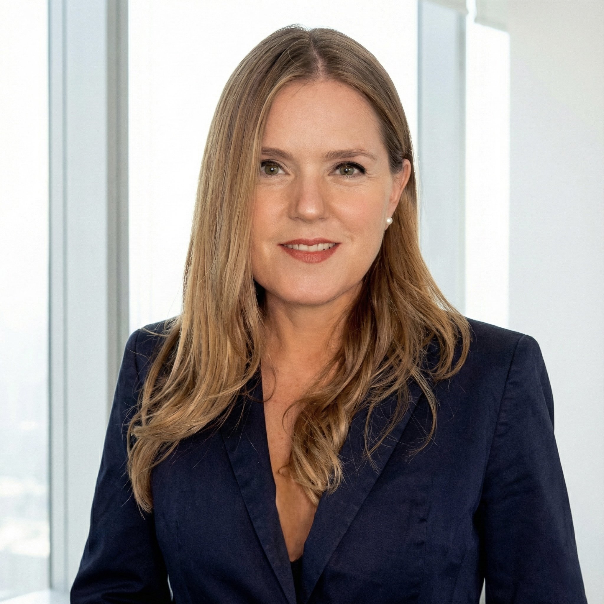 Retrato de una mujer con cabello rubio y liso, sonriendo, vestida con saco azul, en un espacio de oficinas con ventanas altas de fondo.
