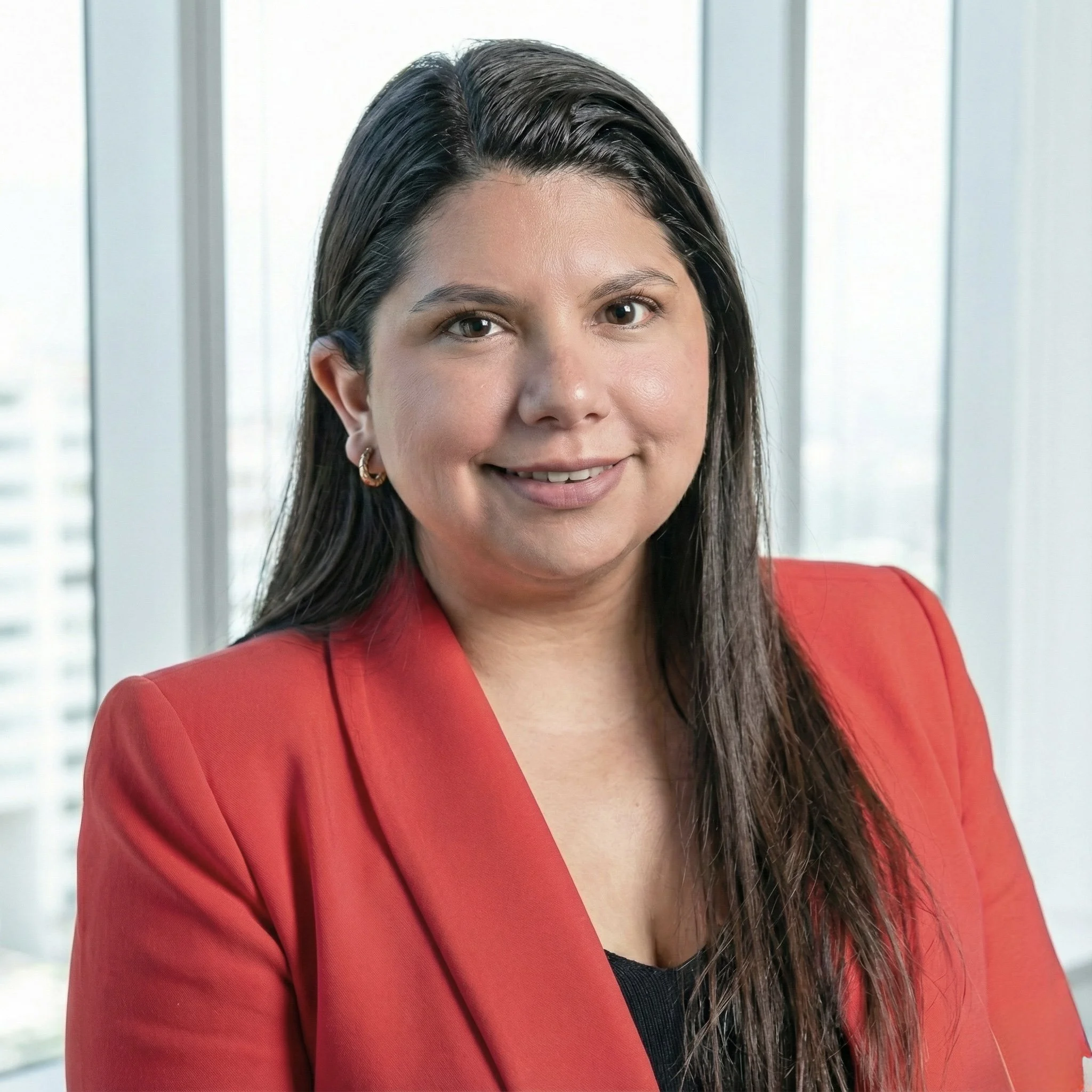 Retrato de una mujer con cabello oscuro y largo, vestida con saco rojo y camiseta negra, posando en una oficina con ventanas y vista de edificios en el fondo.