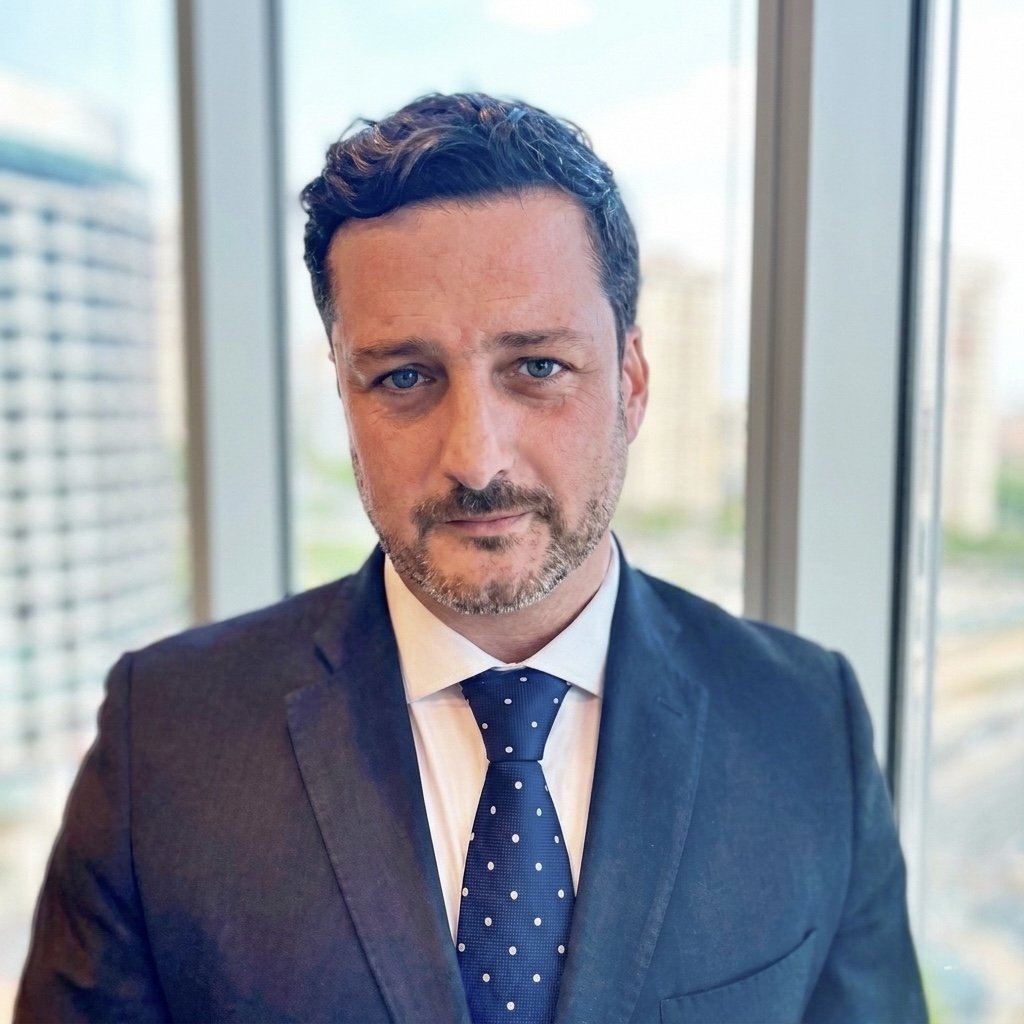 Hombre con barba, cabello oscuro rizado, usando saco oscuro, camisa blanca y corbata azul con lunares blancos, en oficina con ventanas grandes, vista de edificios urbanos al fondo.