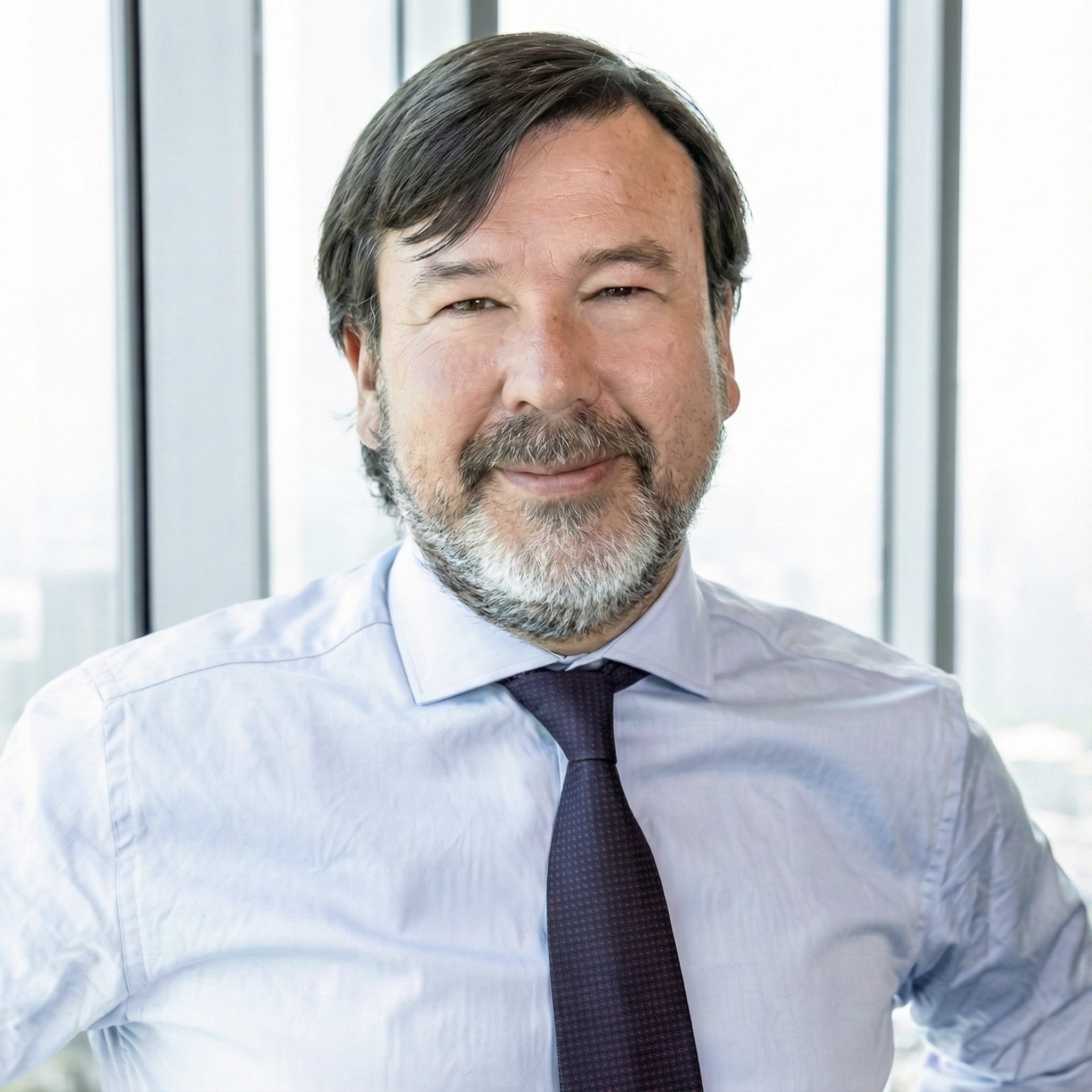 Hombre con barba y cabello castaño, sonriendo, vestido con camisa y corbata en una oficina moderna con ventanas grandes.