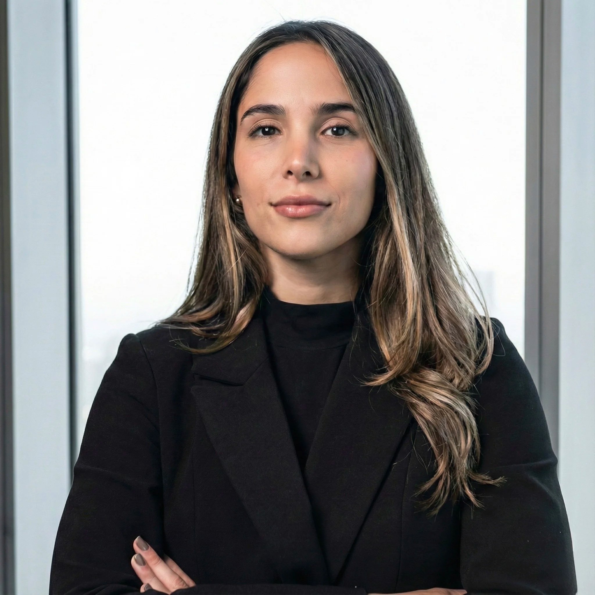 Una mujer con cabello largo y ondulado, vestida con un saco negro, posando con los brazos cruzados en un entorno de oficina con ventanas de fondo.