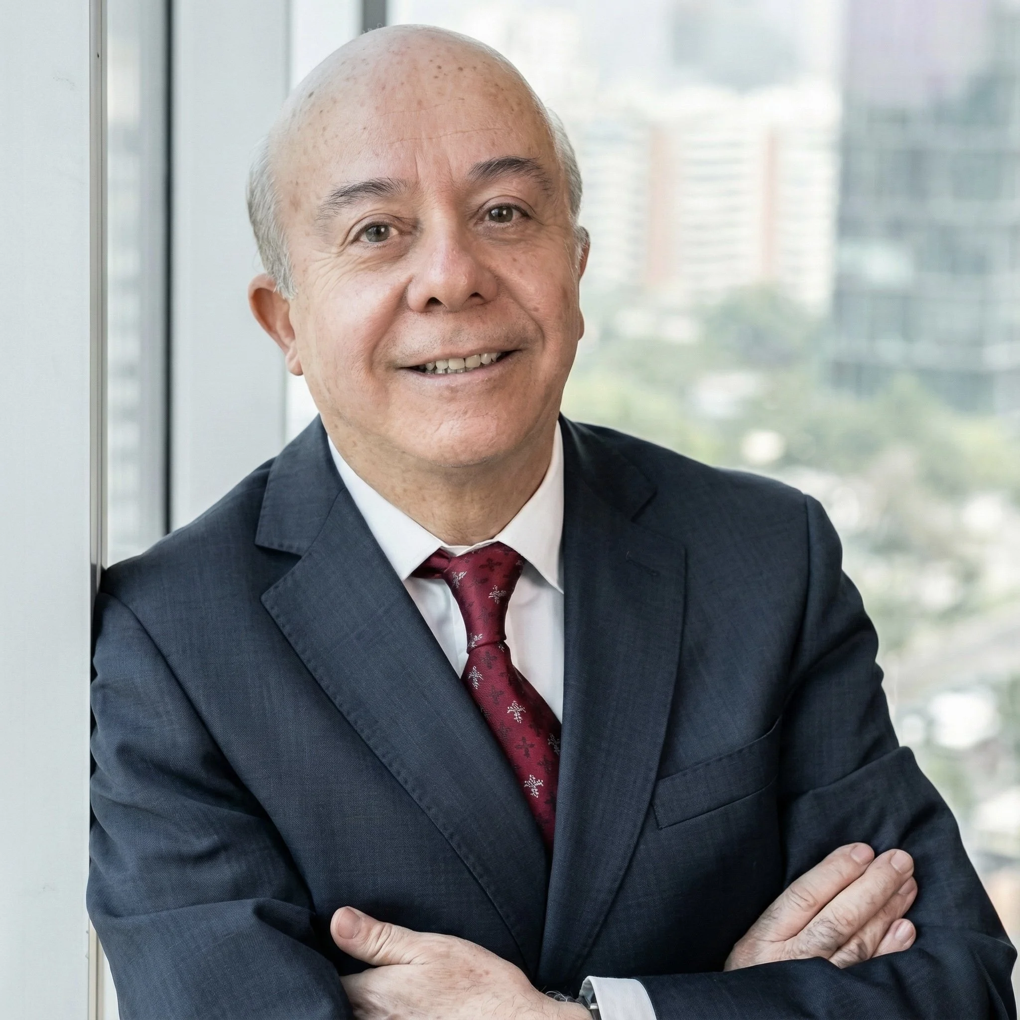 Hombre mayor con traje azul y corbata roja, sonriente, de cabello canoso, en oficina con vista urbana.