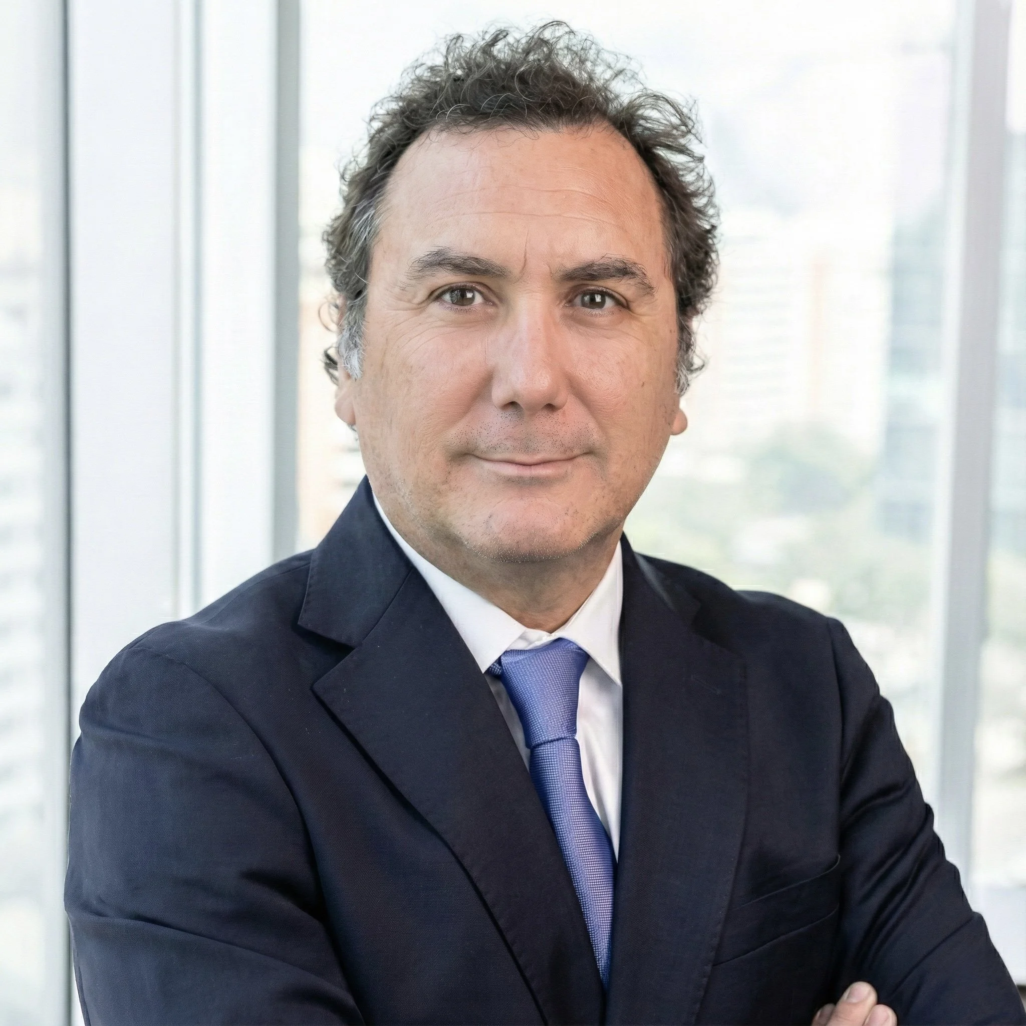 Retrato de un hombre con traje oscuro, camisa blanca y corbata azul, en un ambiente corporativo con ventanas y vista de ciudad de fondo.