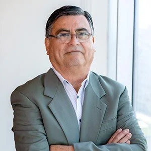 Retrato de un hombre con traje oscuro, camisa blanca y corbata azul, en un ambiente corporativo con ventanas y vista de ciudad de fondo.