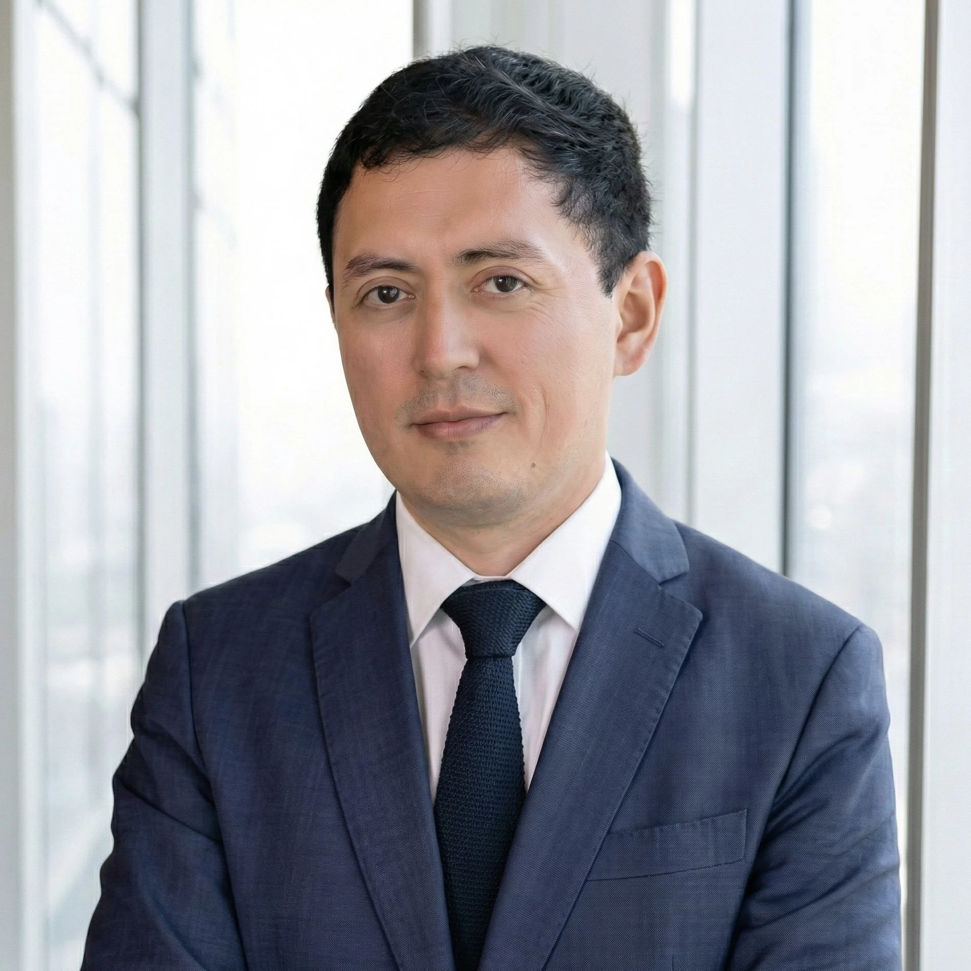 Retrato de un hombre vestido con saco y corbata en un entorno de oficina con ventanas de fondo