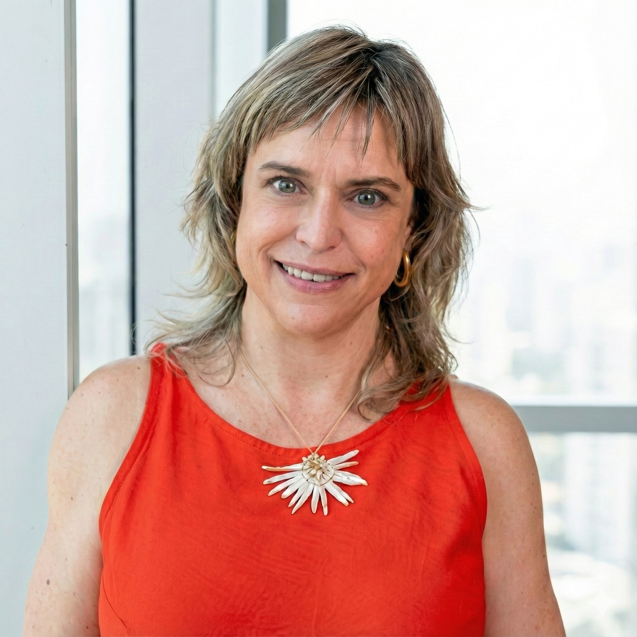 Mujer sonriendo con cabello rubio y ondulado, lleva blusa roja y un collar distintivo en un ambiente claro y moderno.
