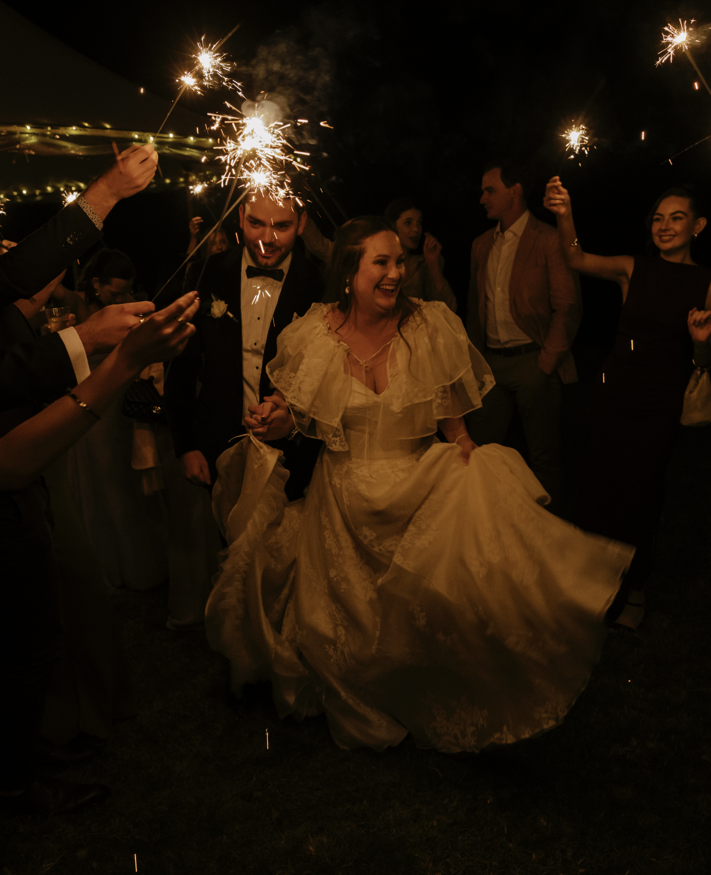 A bride and groom celebrating at night with sparklers, surrounded by friends and family, during a wedding reception.