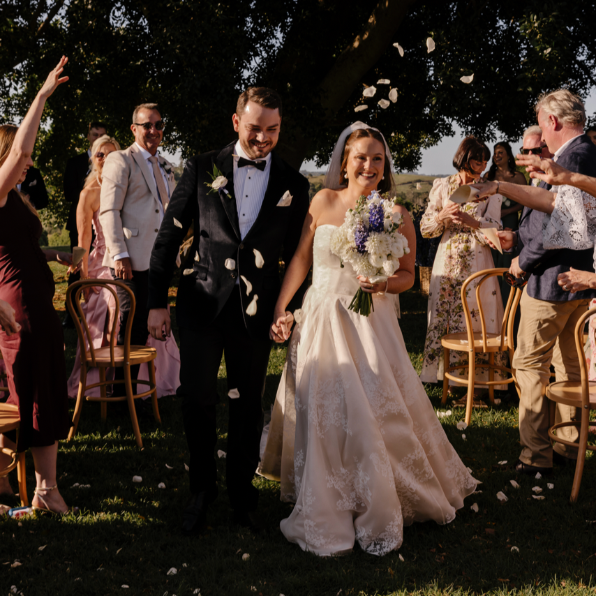 A newly married couple walking hand in hand outdoors, surrounded by friends and family. The bride is wearing a custom Thuy Nguyen Couture wedding dress and holding a bouquet, and the groom is in a black tuxedo.