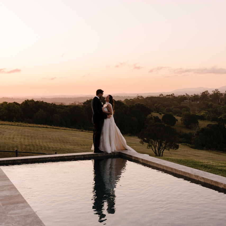 A bride and groom standing close together on the edge of a water feature outdoors at sunset, with a scenic landscape of rolling hills and trees in the background.