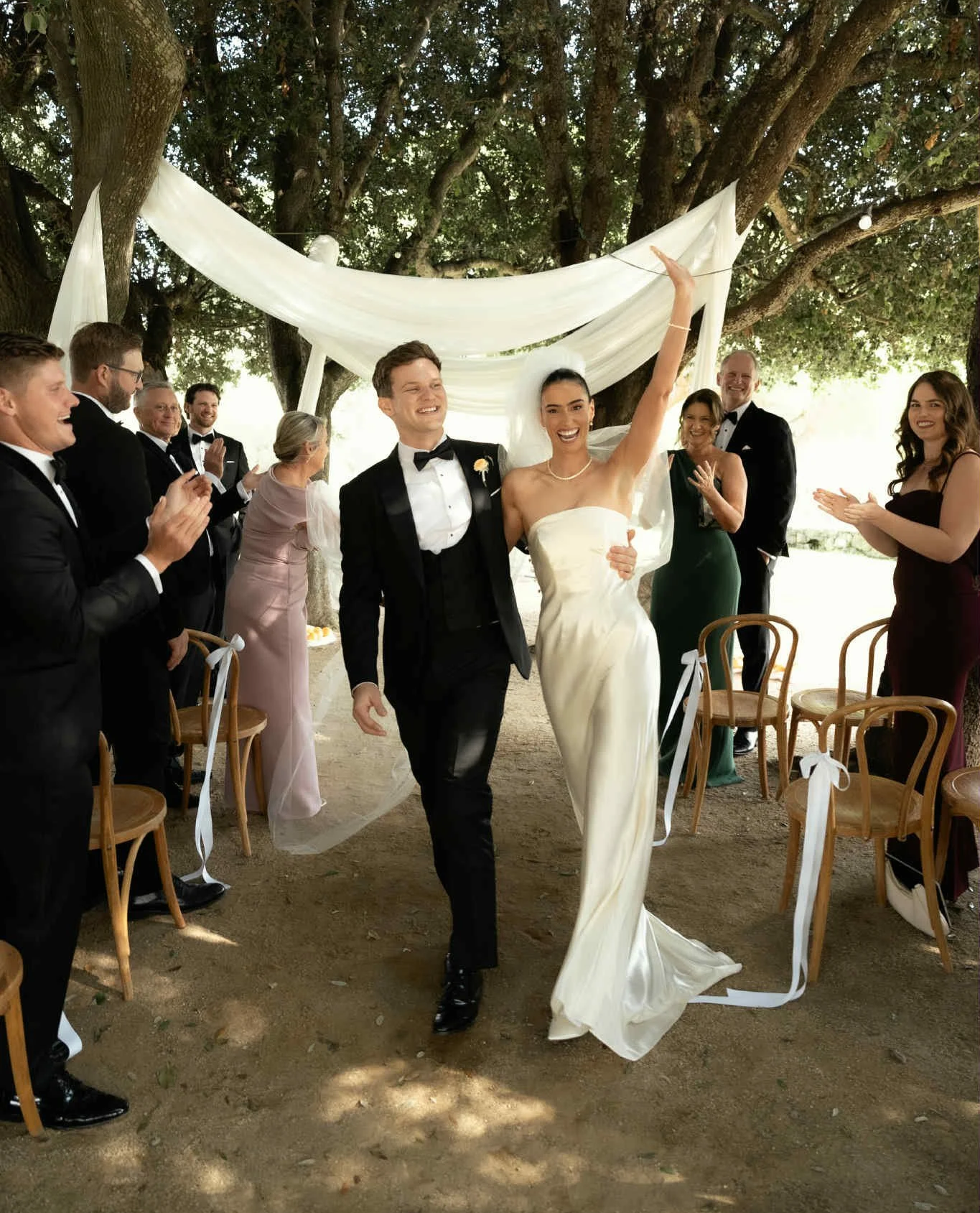 A newly married couple walking down the aisle, smiling, surrounded by clapping friends and family at an outdoor wedding under trees with a white fabric canopy.