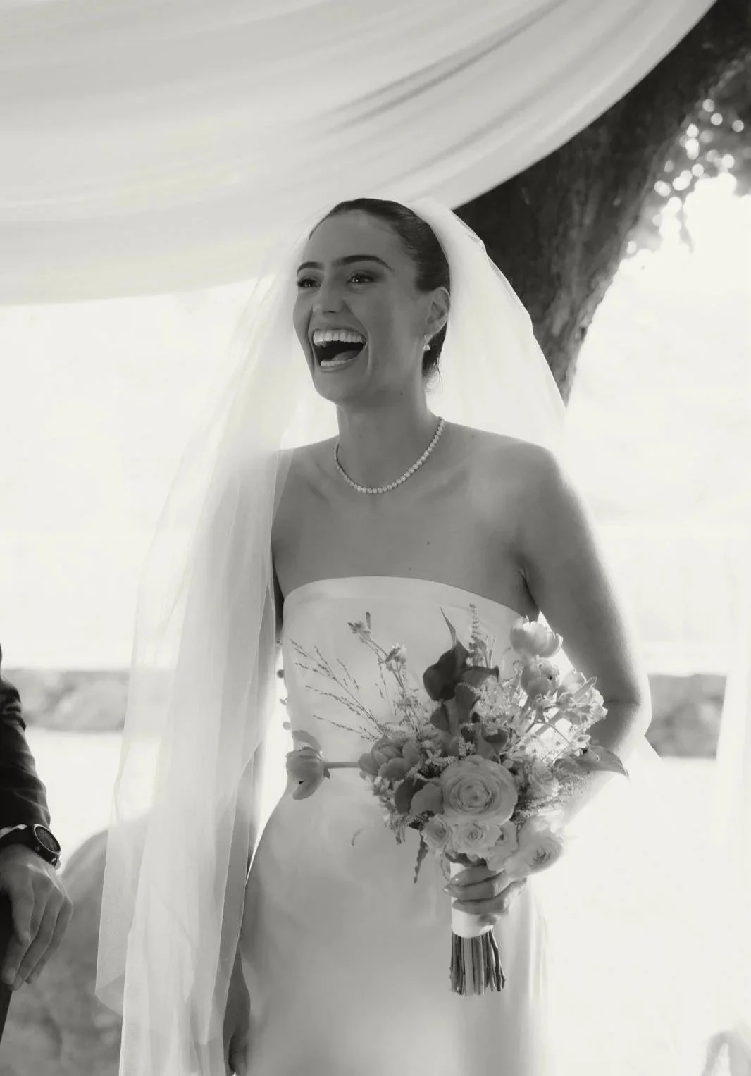 Black and white photo of a smiling bride in a strapless wedding dress by Thuy Nguyen Couture holding a bouquet of flowers, wearing a veil and pearl necklace, standing outdoors near a tree.