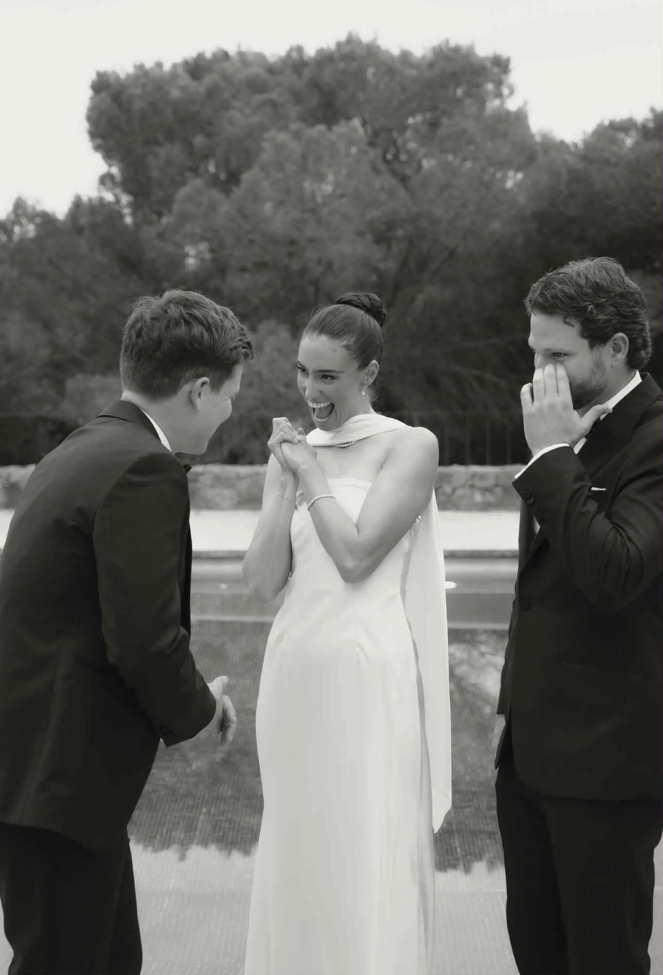 A woman in a wedding dress is happily reacting while a man in a suit proposes to her. Another man in a suit is laughing and covering his mouth. They are outdoors with trees in the background. Bespoke Bridal Brisbane.