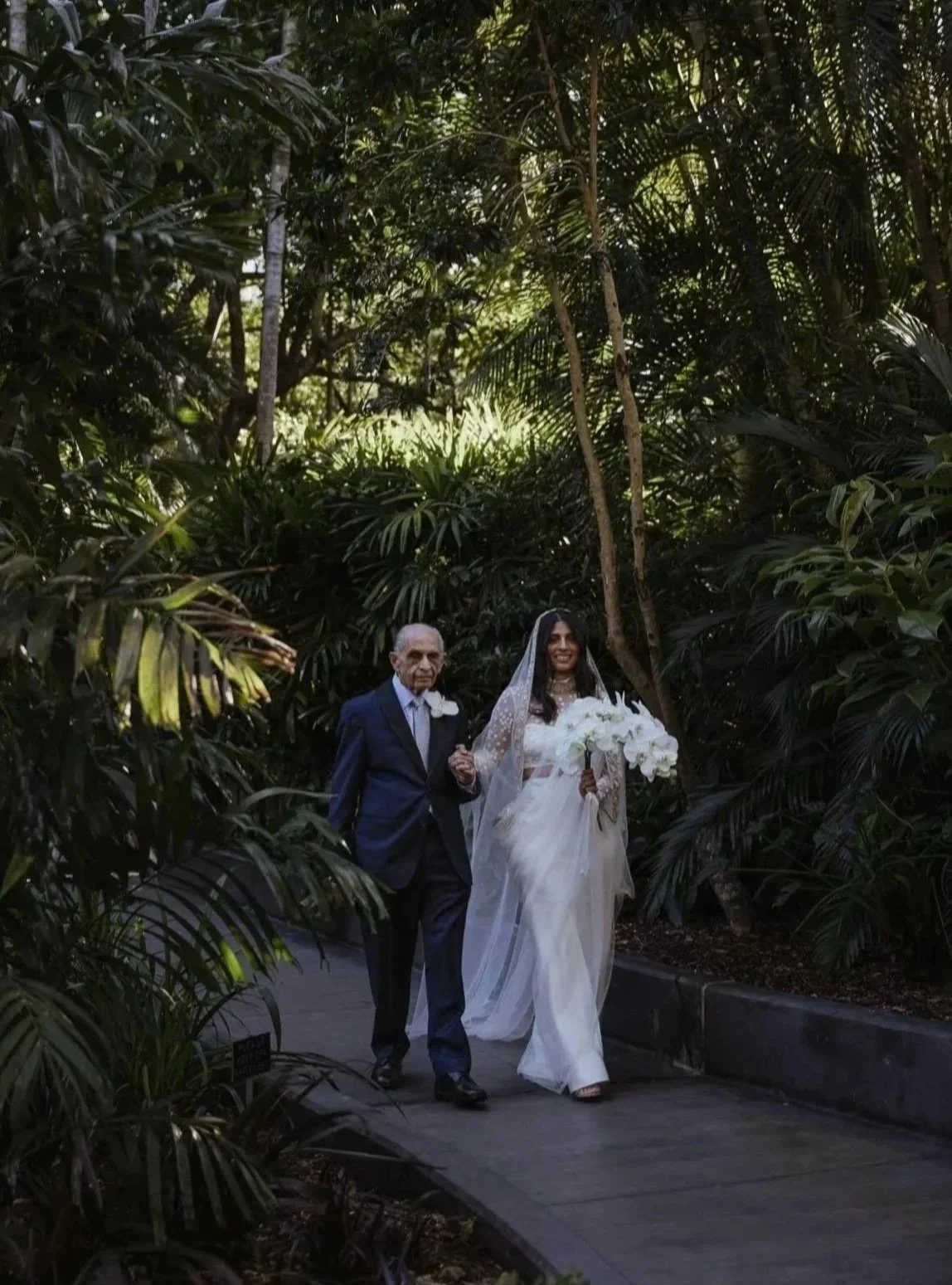 A bride in a white wedding dress holding a bouquet, walking on a path beside an older man in a dark suit, surrounded by lush green jungle vegetation.