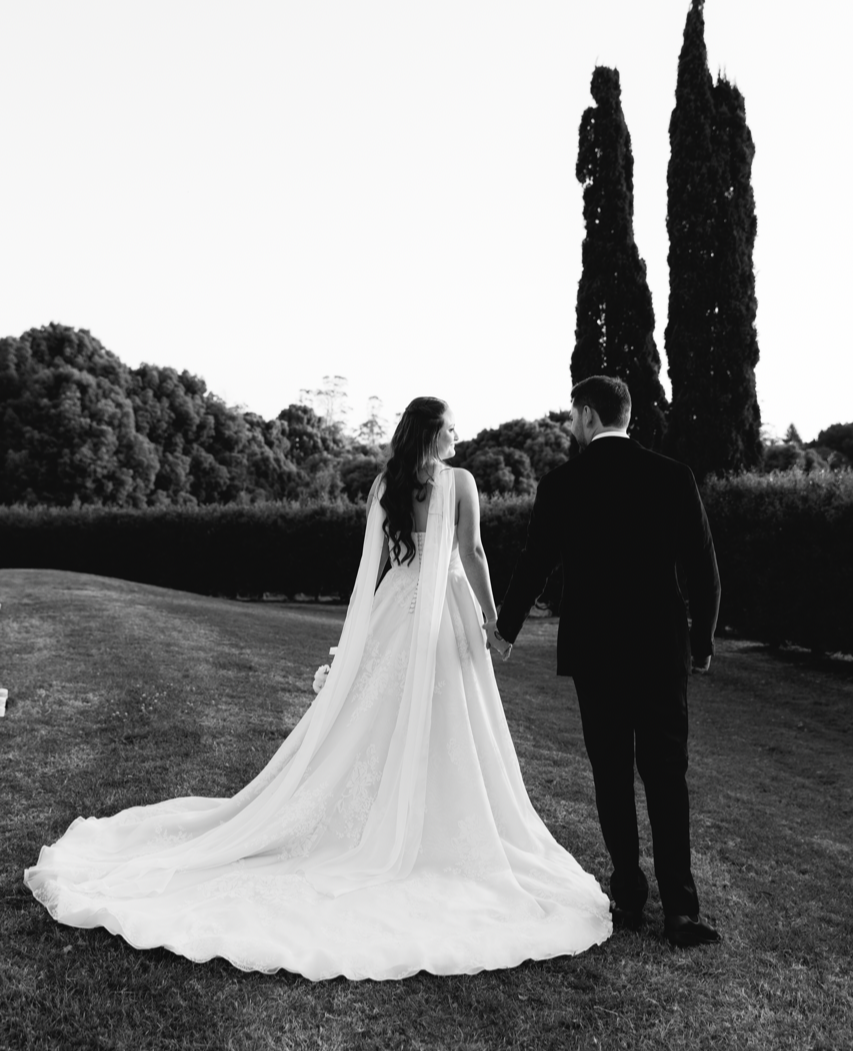Black and white photograph of a bride and groom walking hand in hand outdoors, with tall trees and a clear sky in the background. Bride is wearing a custom Thuy Nguyen Couture wedding dress with a scarf.