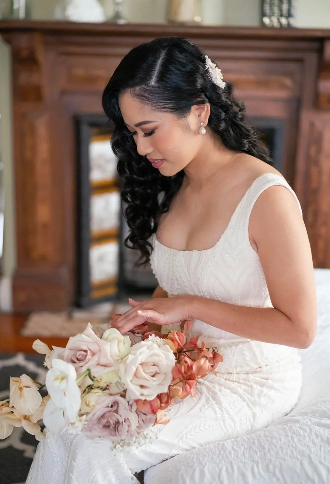 A bride sitting on a bed holding a bouquet of pink and white roses, dressed in a white wedding gown with lace details, with dark curly hair adorned with a white flower accessory, in a room with wooden furniture.