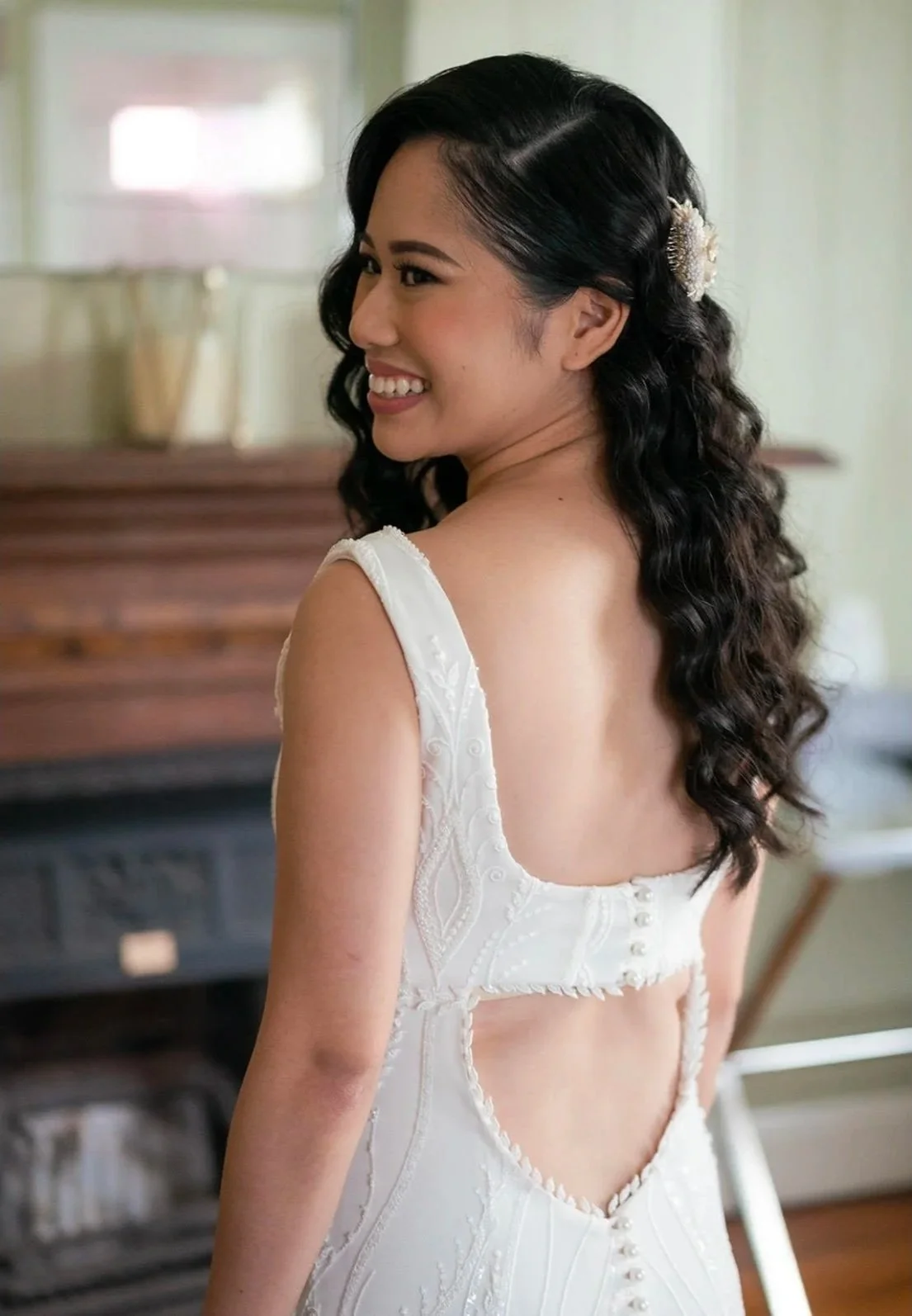 A woman with long, dark, curly hair smiling, wearing a white backless dress with embroidered details and buttons, in a room with a piano in the background.