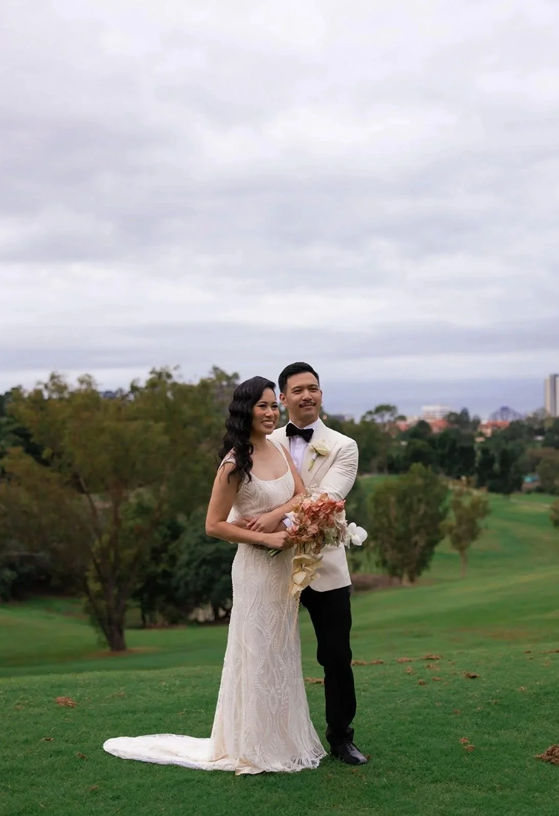 A bride and groom stand together outdoors on a grassy field, smiling. The bride wears a white lace wedding gown and holds a bouquet of pink and white flowers. The groom wears a white tuxedo jacket with black pants and a black bow tie. The background 