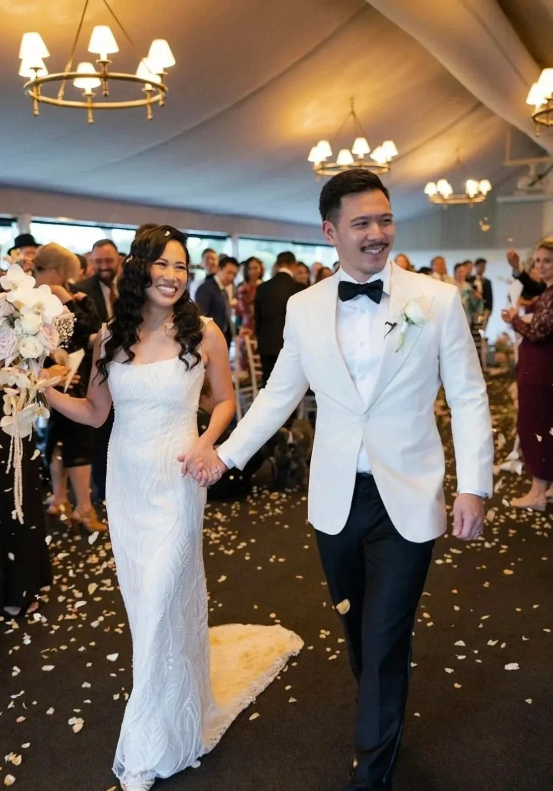 A newly married couple walking down the aisle during their wedding reception, holding hands and smiling, with guests clapping and celebrating in the background.