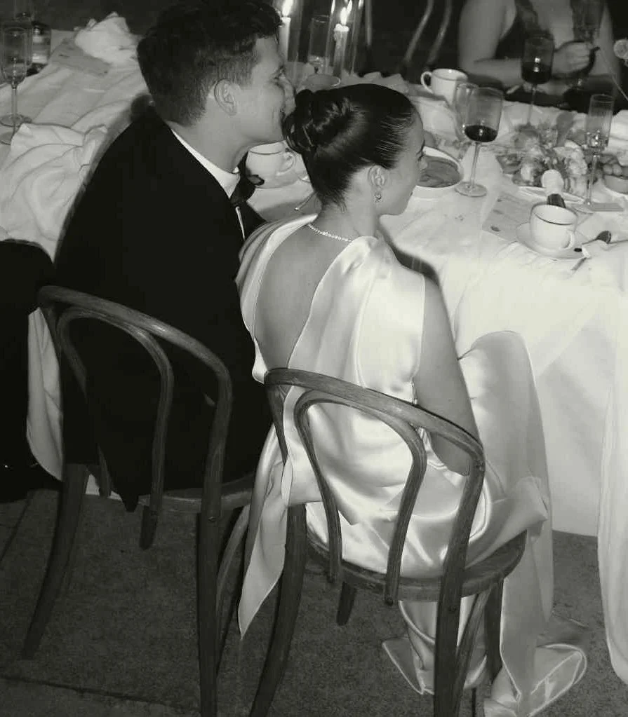 A black-and-white photo of a man and woman sitting at a table during a formal event. They are dressed elegantly, with the woman wearing a satin gown and pearl necklace, and the man in a tuxedo. The table has glasses, cups, and food items. The woman h