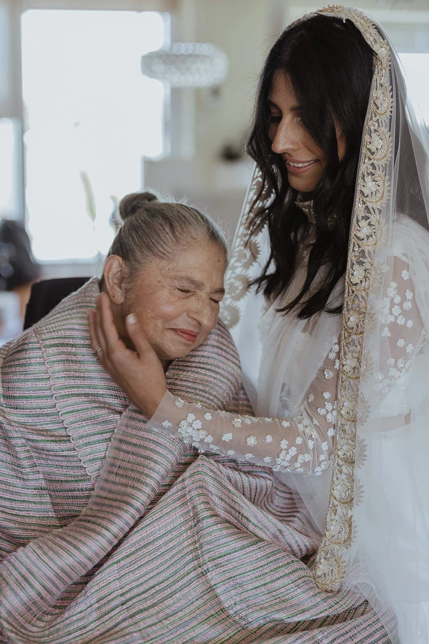 A woman in a wedding dress gently holds and comforts an elderly woman with closed eyes, sitting in a bright room.