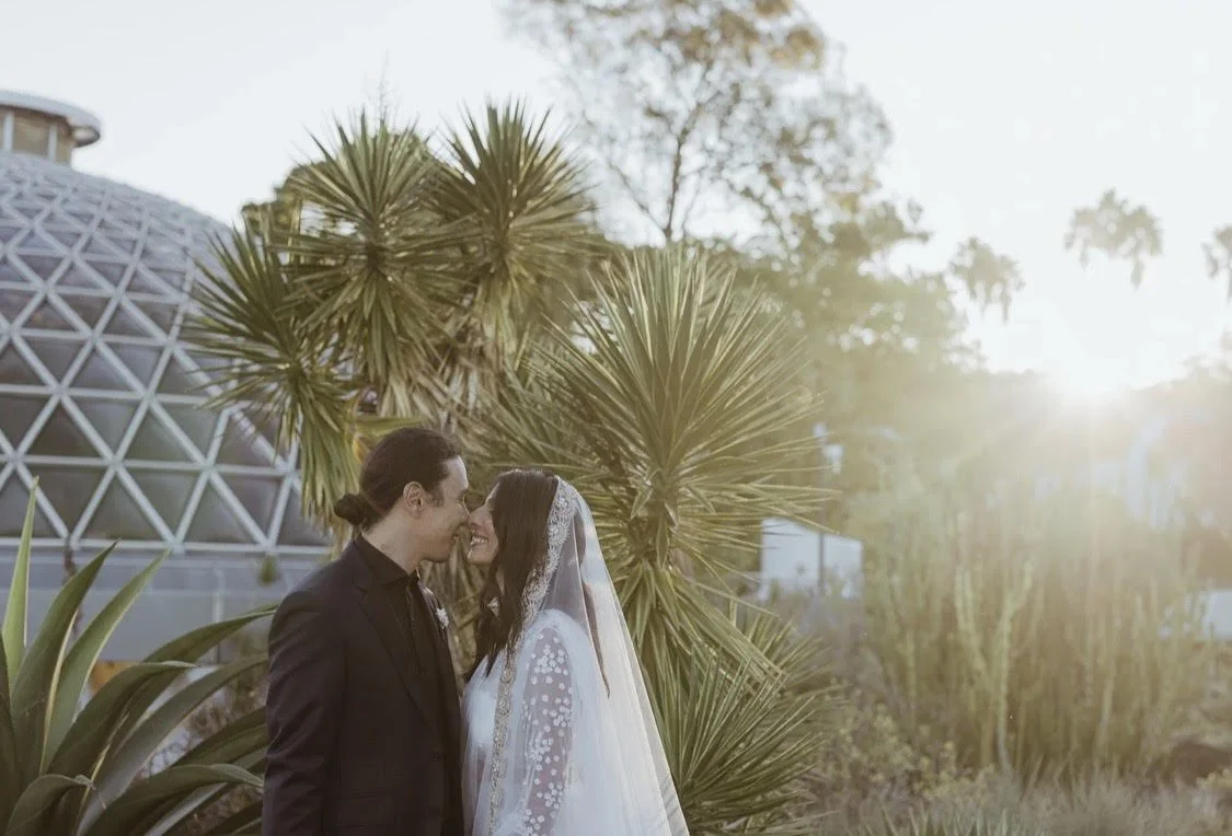 A bride and groom stand close, touching foreheads and smiling at each other outdoors during sunset, with tropical plants behind them.