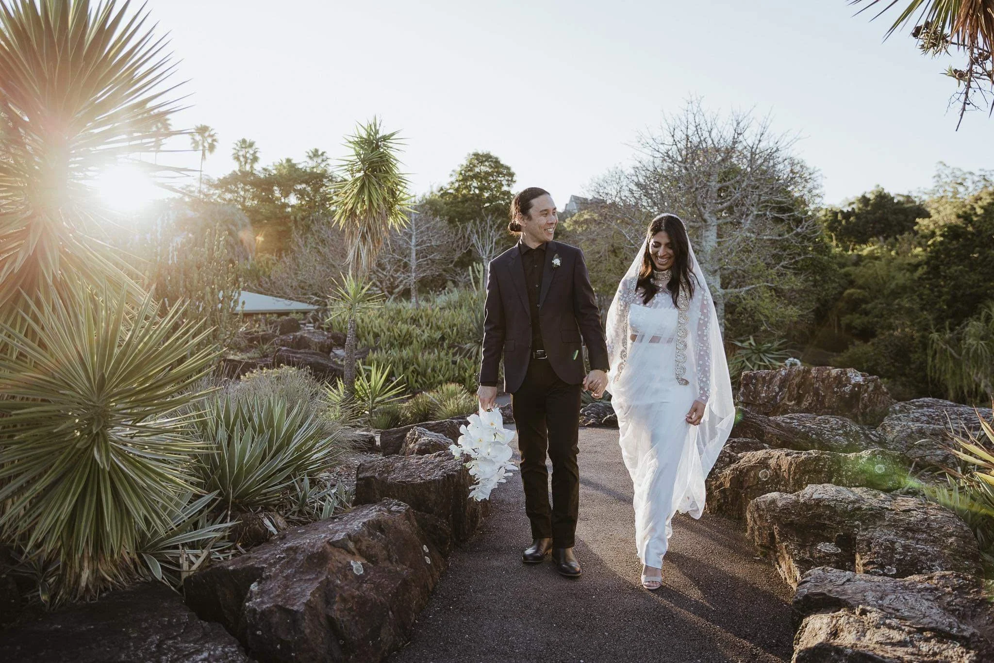 A newlywed couple walking hand in hand through a garden path surrounded by desert plants, with the sun setting behind them, smiling and happily looking at each other.