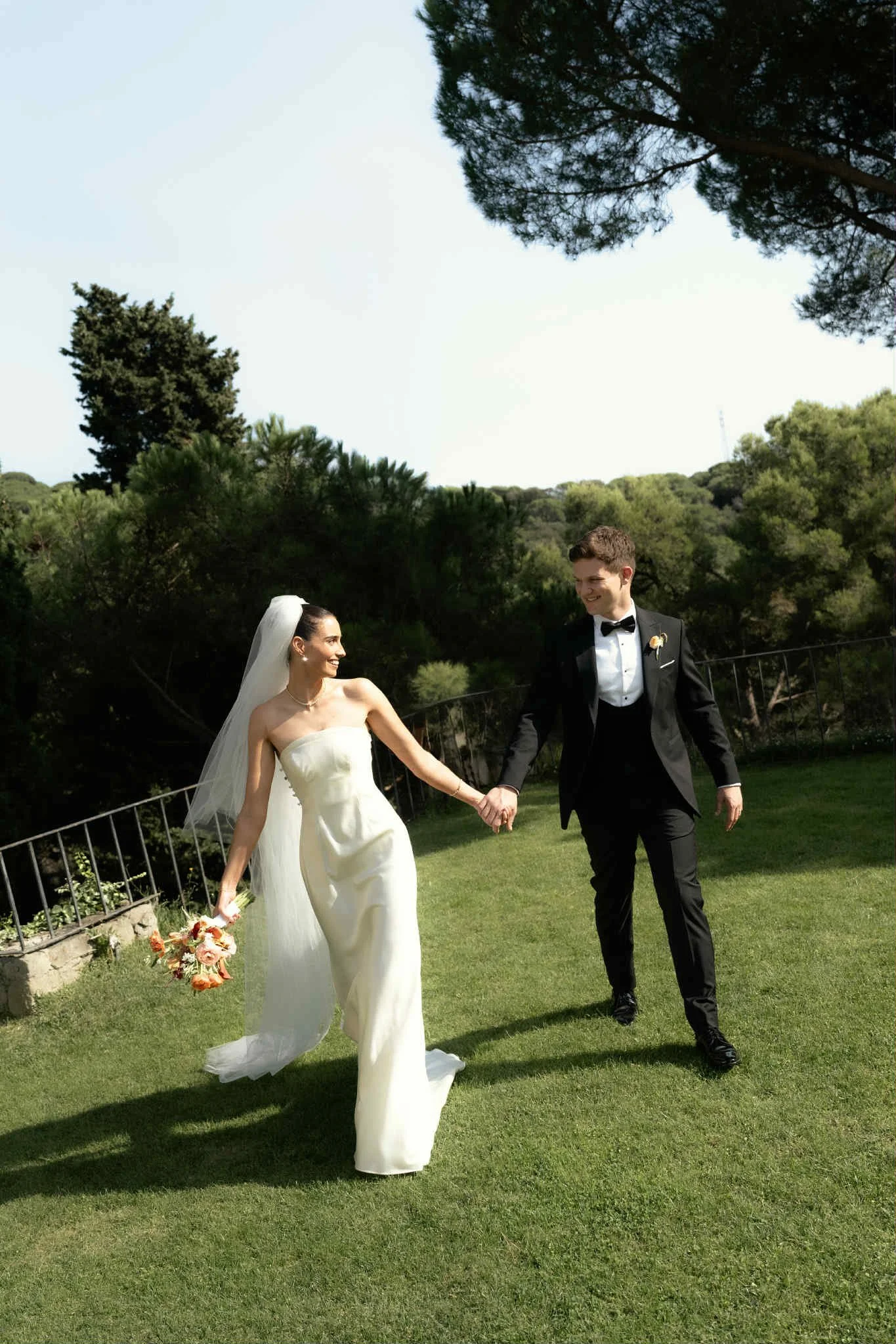 A bride and groom holding hands and walking on a grassy area during their wedding, with trees and a metal fence in the background.