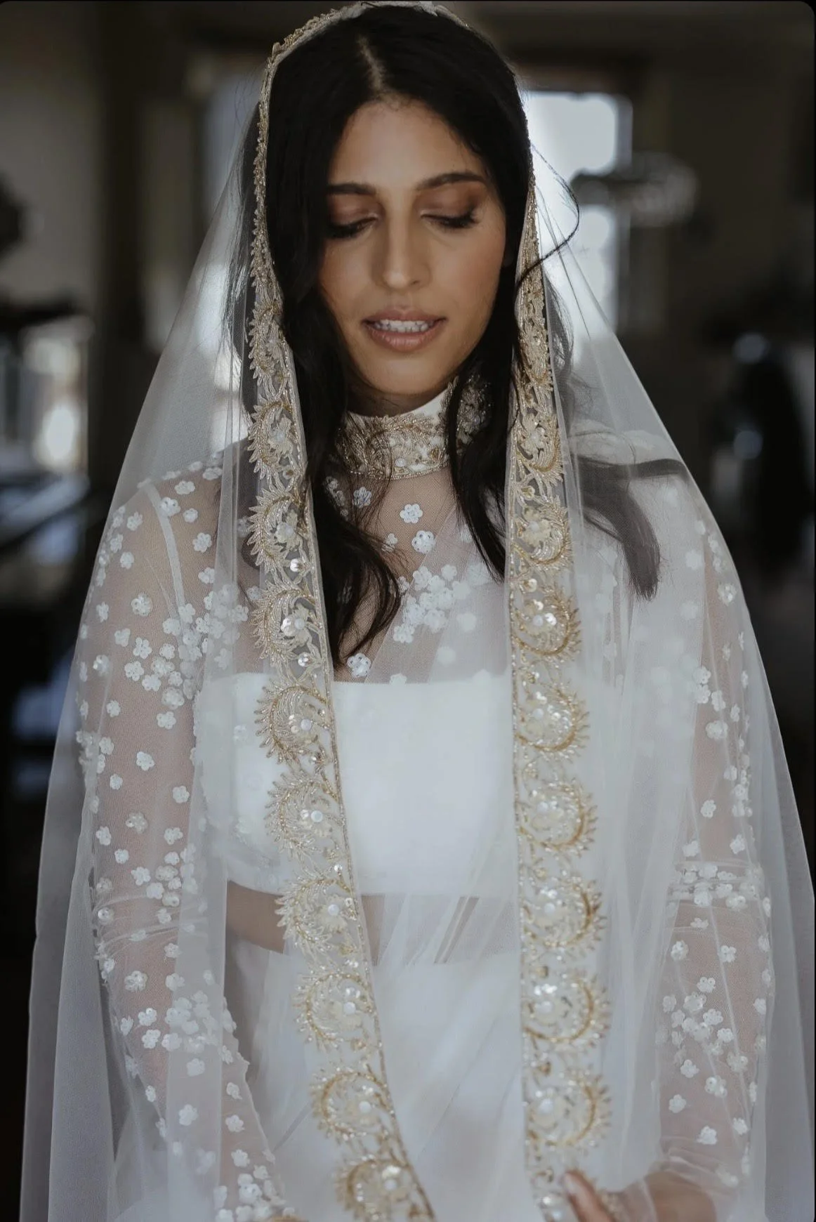 A woman with dark hair and a bridal veil, looking down, wearing a white wedding dress designed and made by Thuy Nguyen Couture with floral embroidery and gold embroidered trim. Bespoke Bridal Brisbane.
