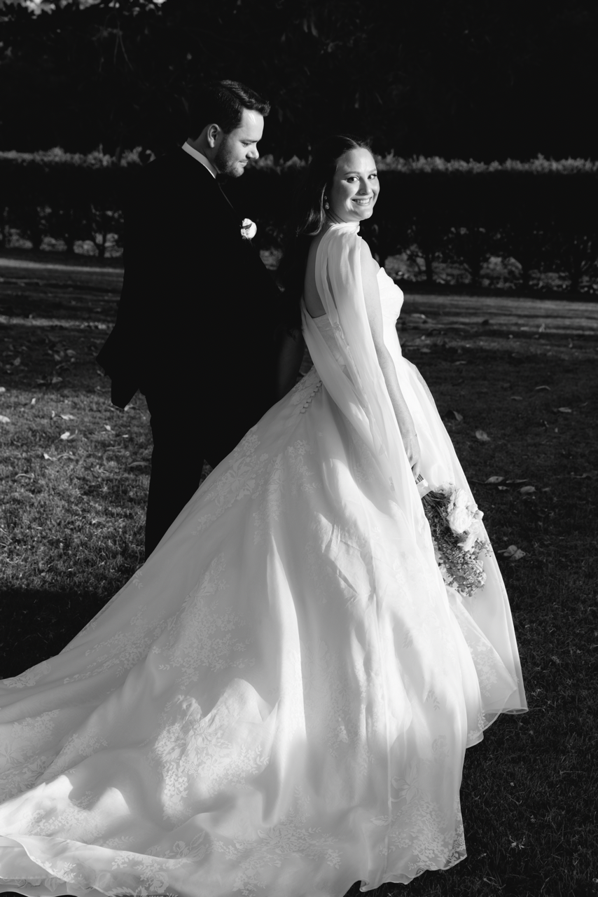 A black and white photo of a bride wearing a custom Thuy Nguyen Couture wedding dress holding a bouquet, standing beside a groom in a tuxedo, both outdoors at night.