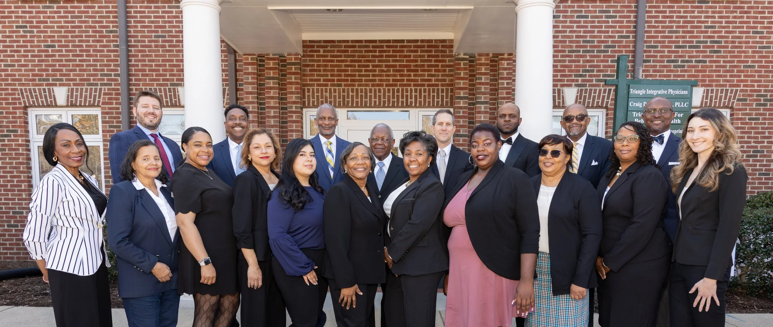 Group of professionally dressed men and women standing outside a brick building, smiling for a photo.
