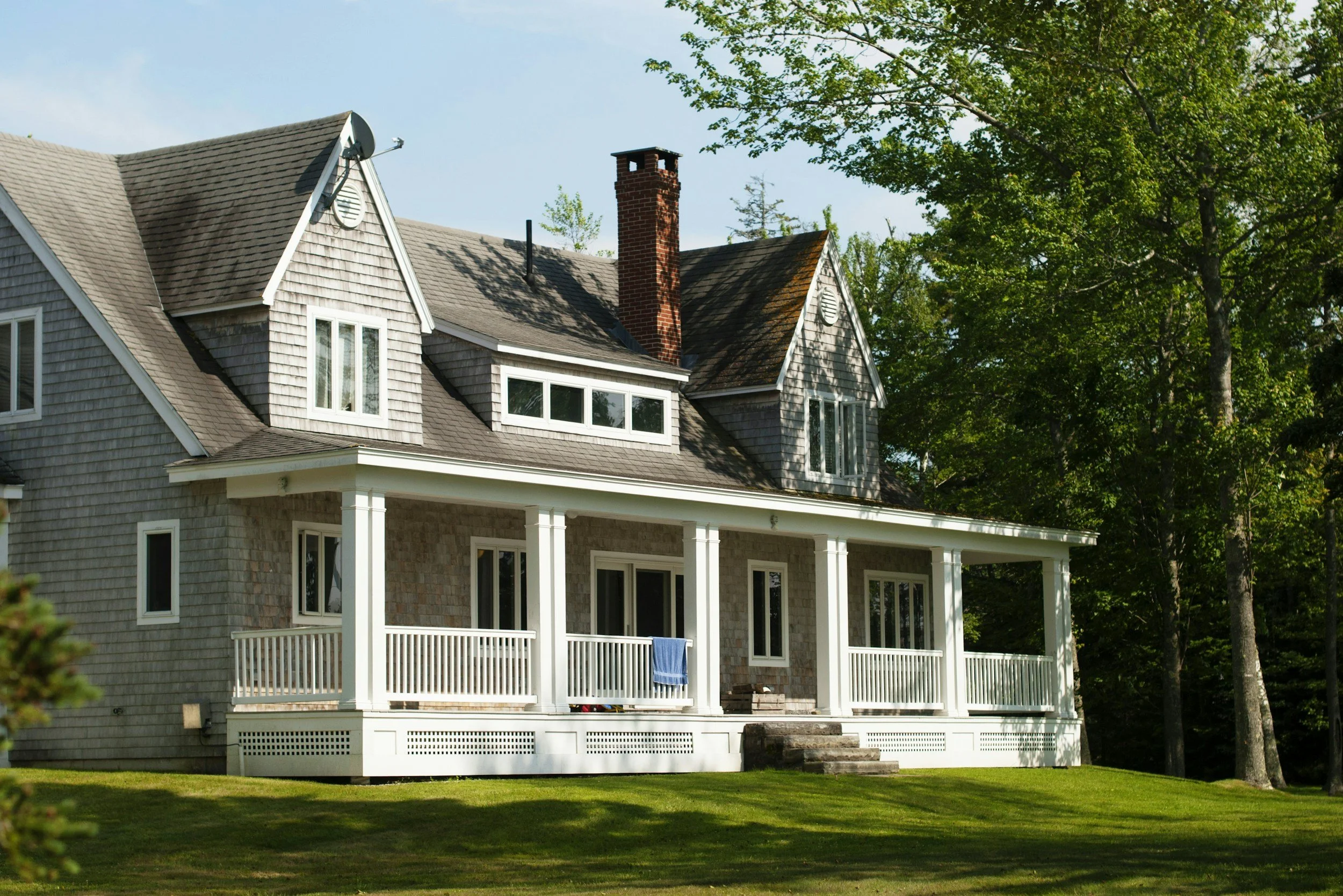 A large house with a front porch, multiple windows, gray shingle siding, and a chimney surrounded by tall trees and a grassy yard.