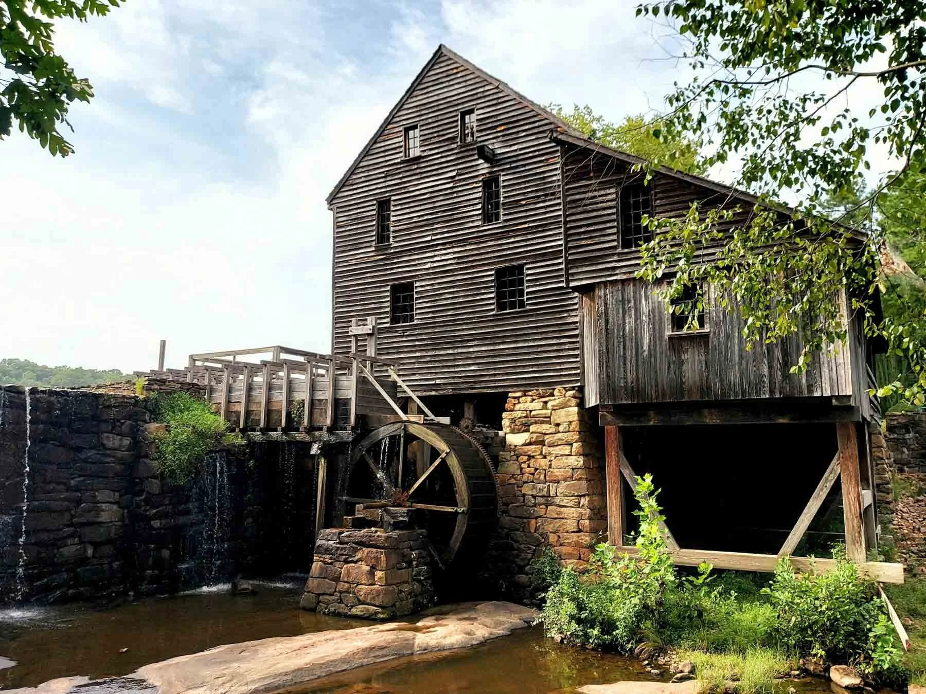 Historic Yates Mill Raleigh NC: Old wooden gristmill with water wheel beside a creek, surrounded by trees.