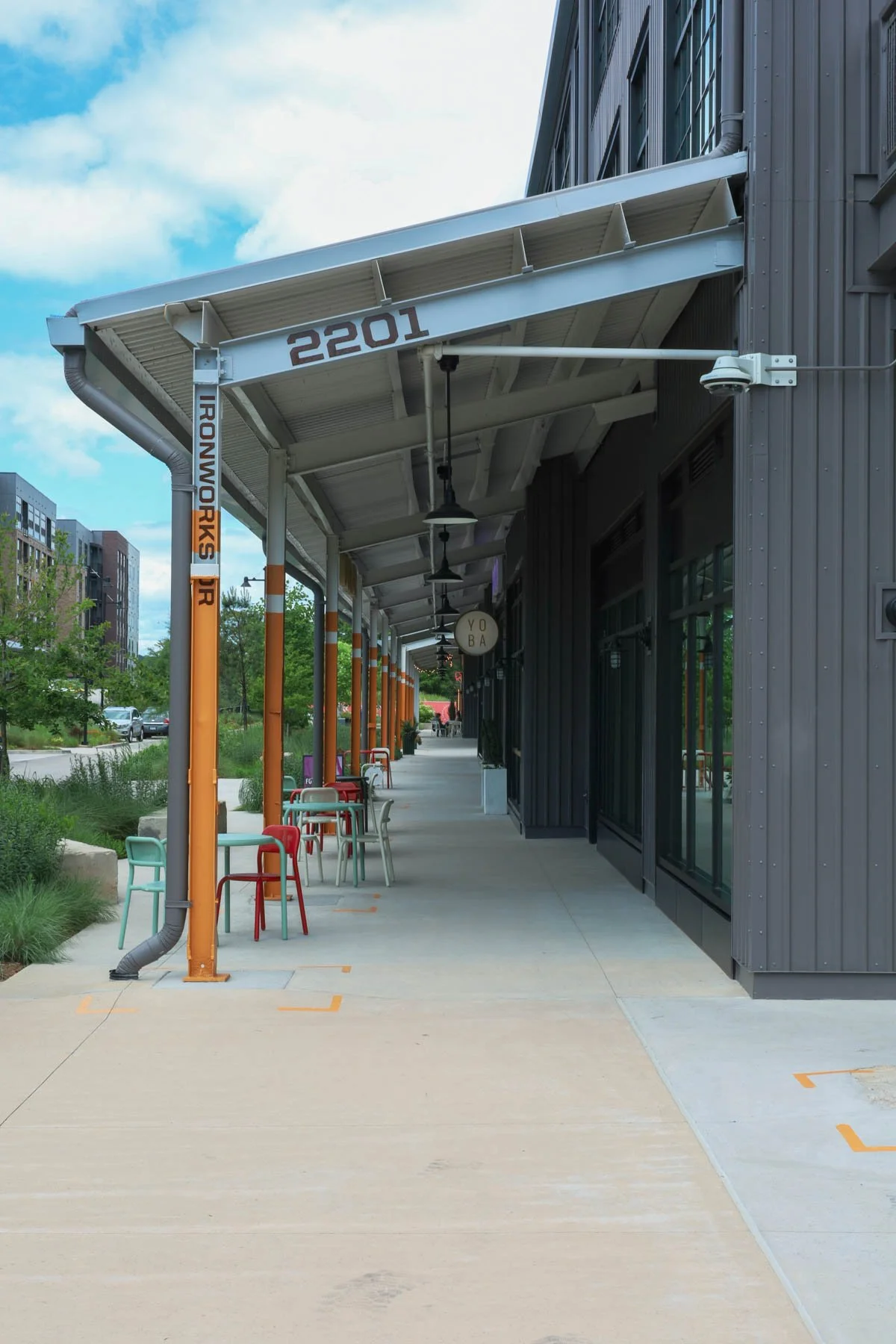 Empty sidewalk outside a modern building with colorful chairs, metal roof, and signage reading 'IRWONWORKS' and the number 2201.