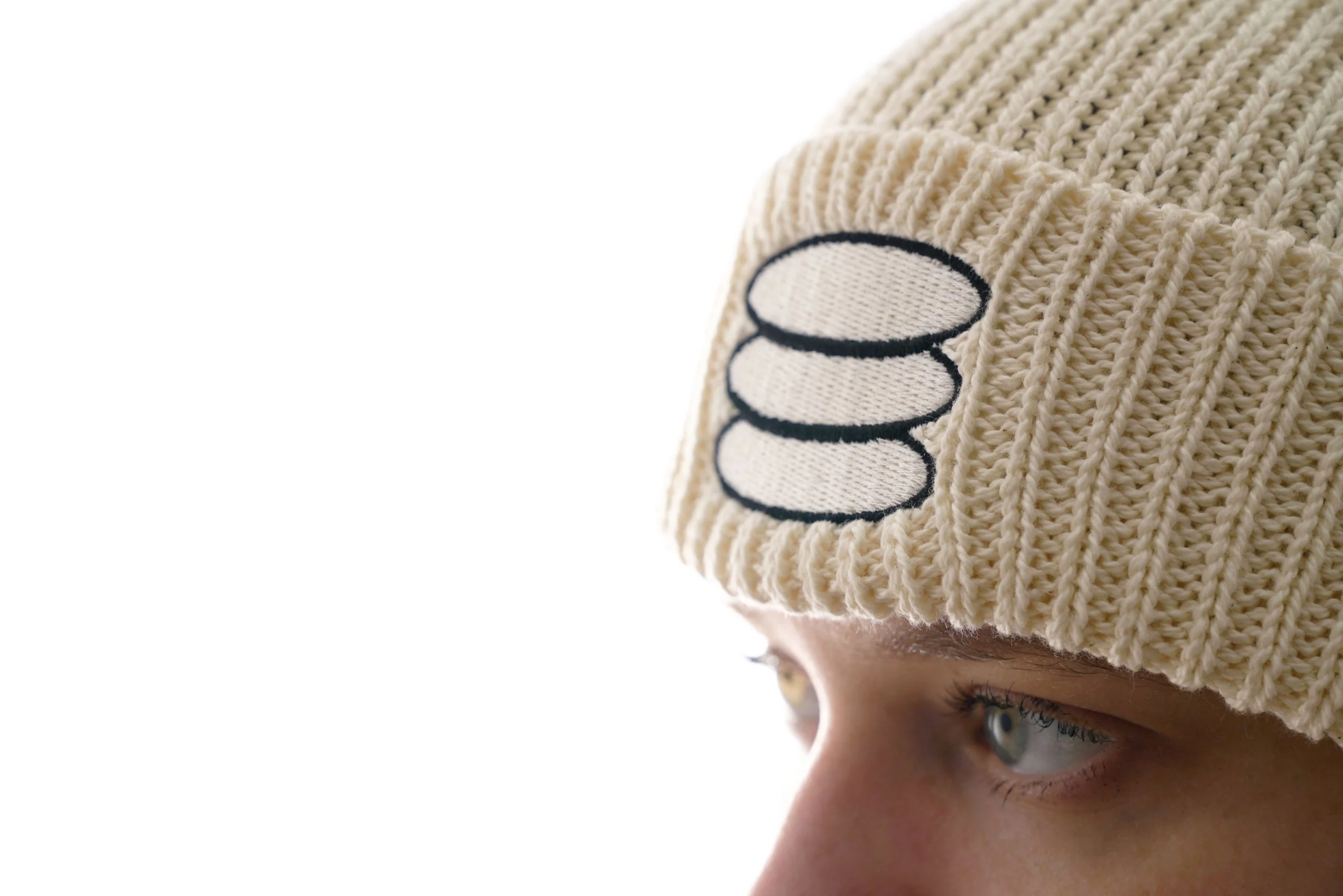 Close-up of a person wearing a beige knit beanie with a black and white embroidered patch, showing only part of their face and their blue eyes.