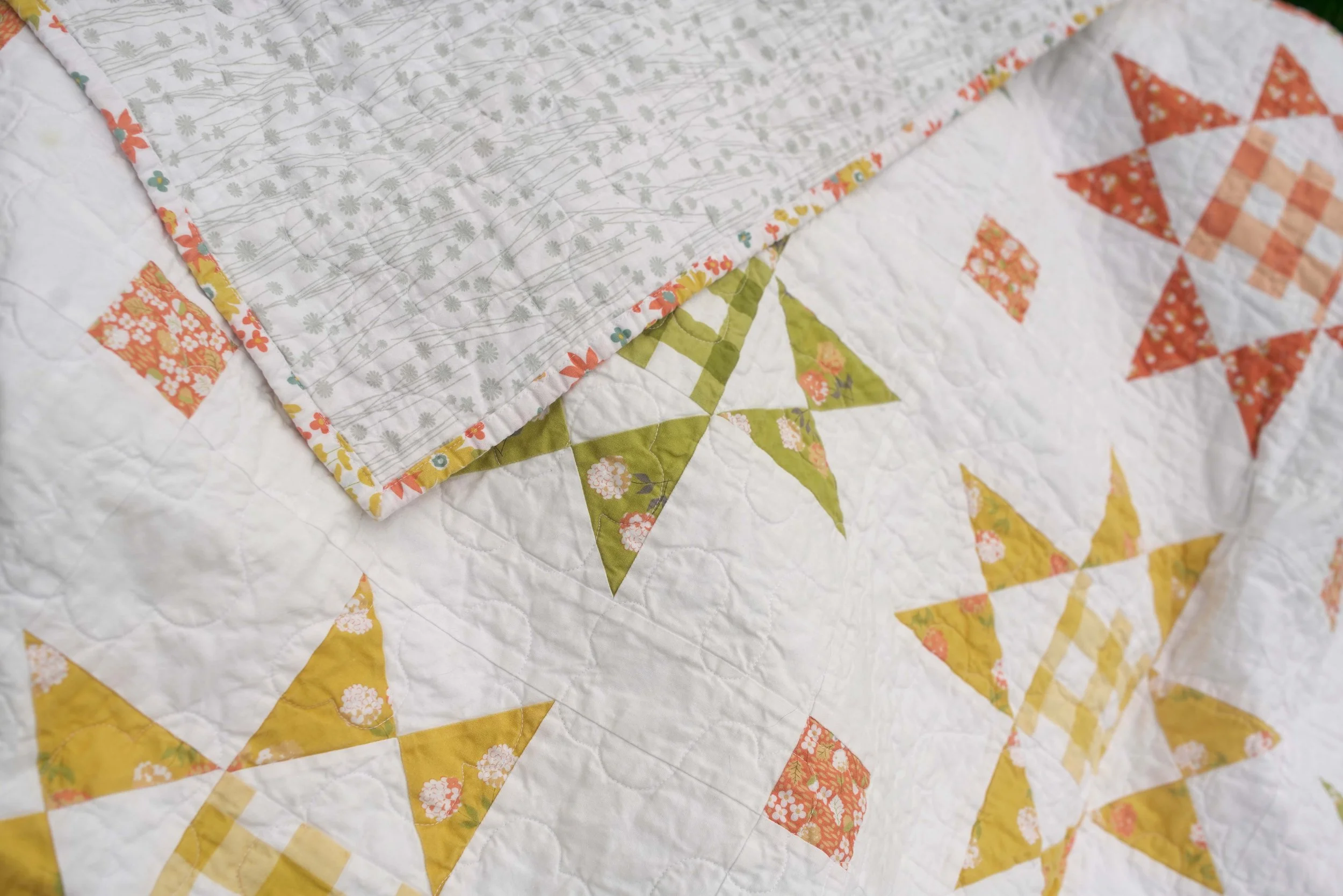Close-up of a quilt with star patterns in yellow, orange, and green, featuring floral designs, and a layered fabric with a gray floral print on top.