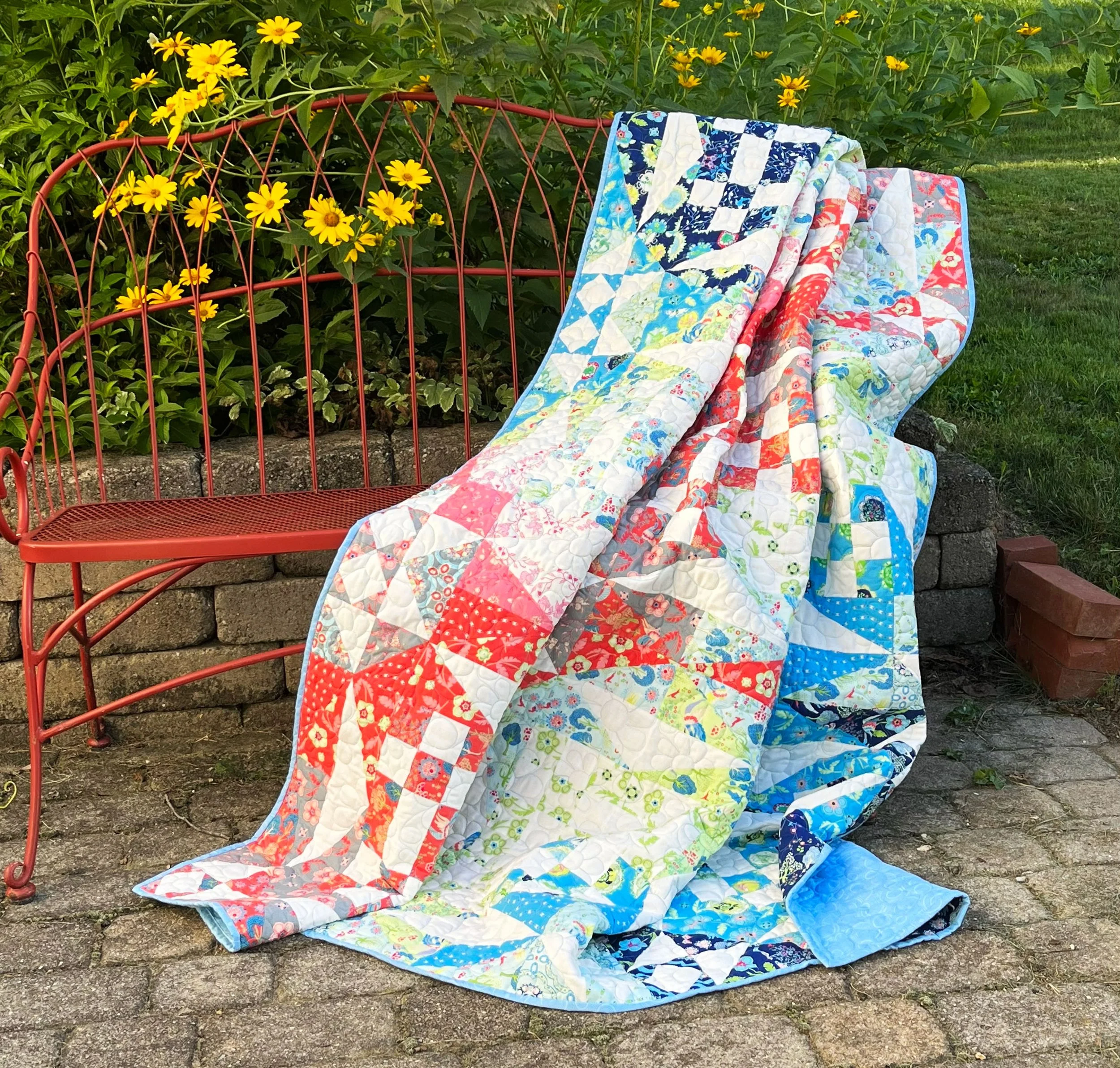 A colorful quilt draped over a red metal garden bench outdoors with yellow flowers and green foliage in the background.