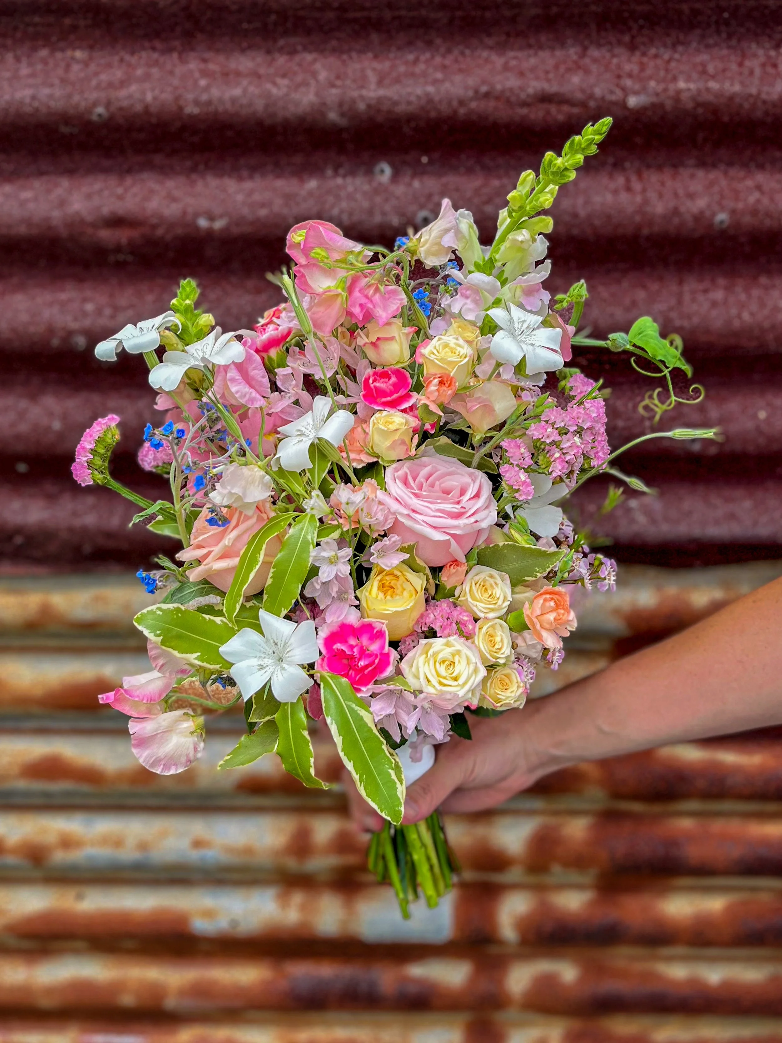 A colorful, whimsical bridal bouquet of mixed flowers in front of a rustic tin wall.