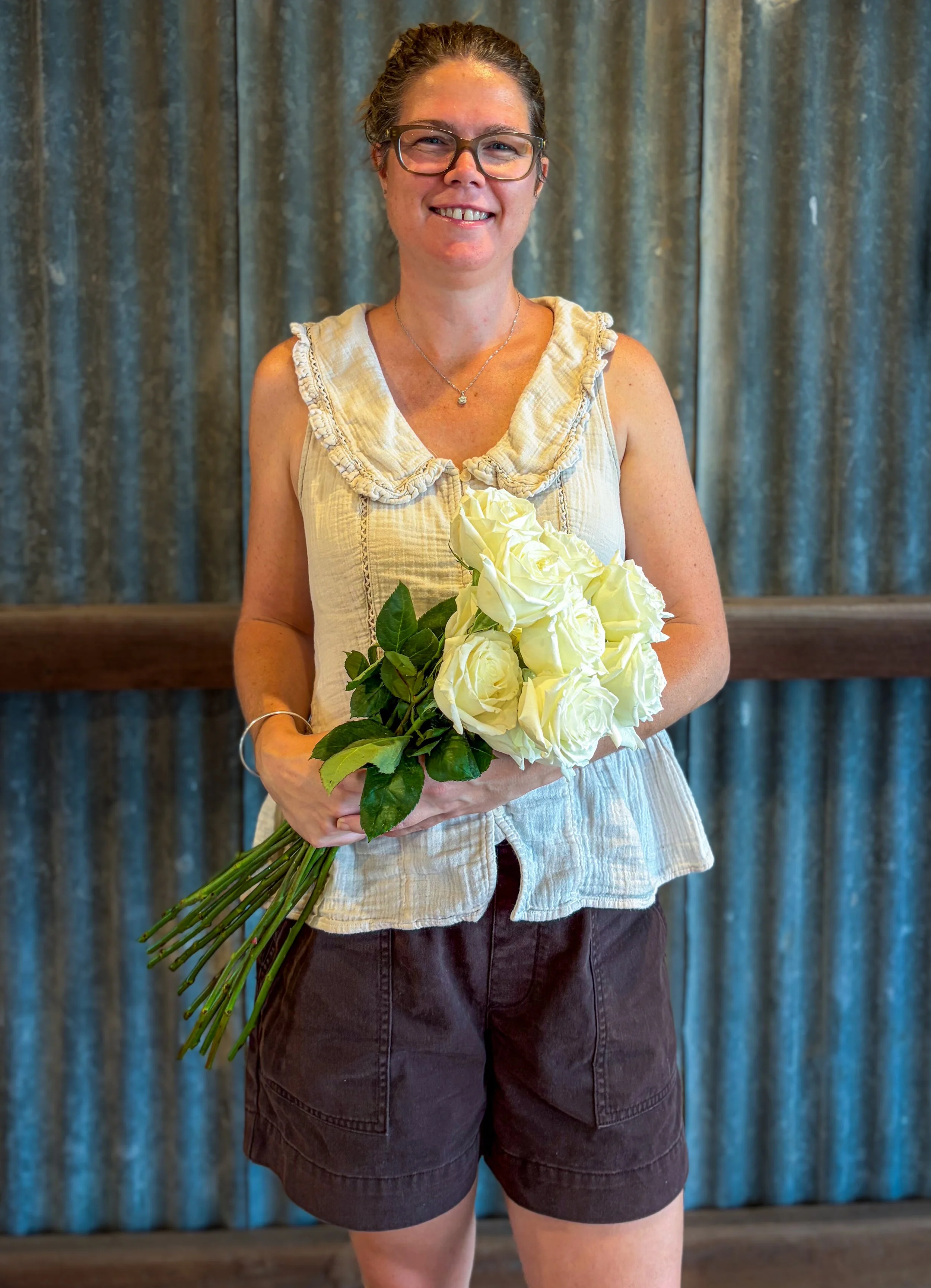 A florist holding a bouquet of white roses, standing in front of a textured rusted tin background, smiling at the camera.