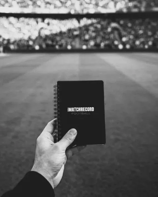 A person's hand holding a black notebook labeled "MATCH RECORD FOOTBALL" on a stadium field with empty seats in the background.