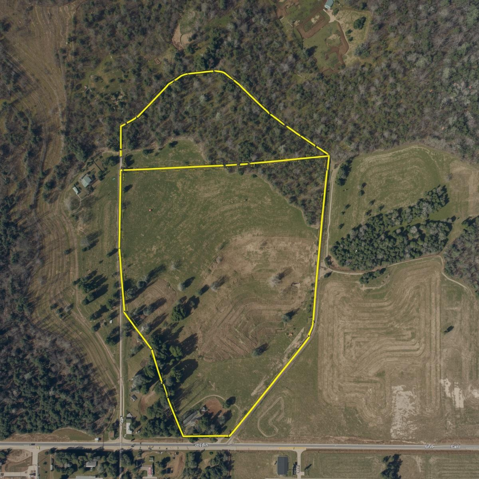 Aerial view of a large, irregularly shaped plot of land outlined in yellow, with open fields, wooded areas, and a few small structures, adjacent to a road at the bottom.