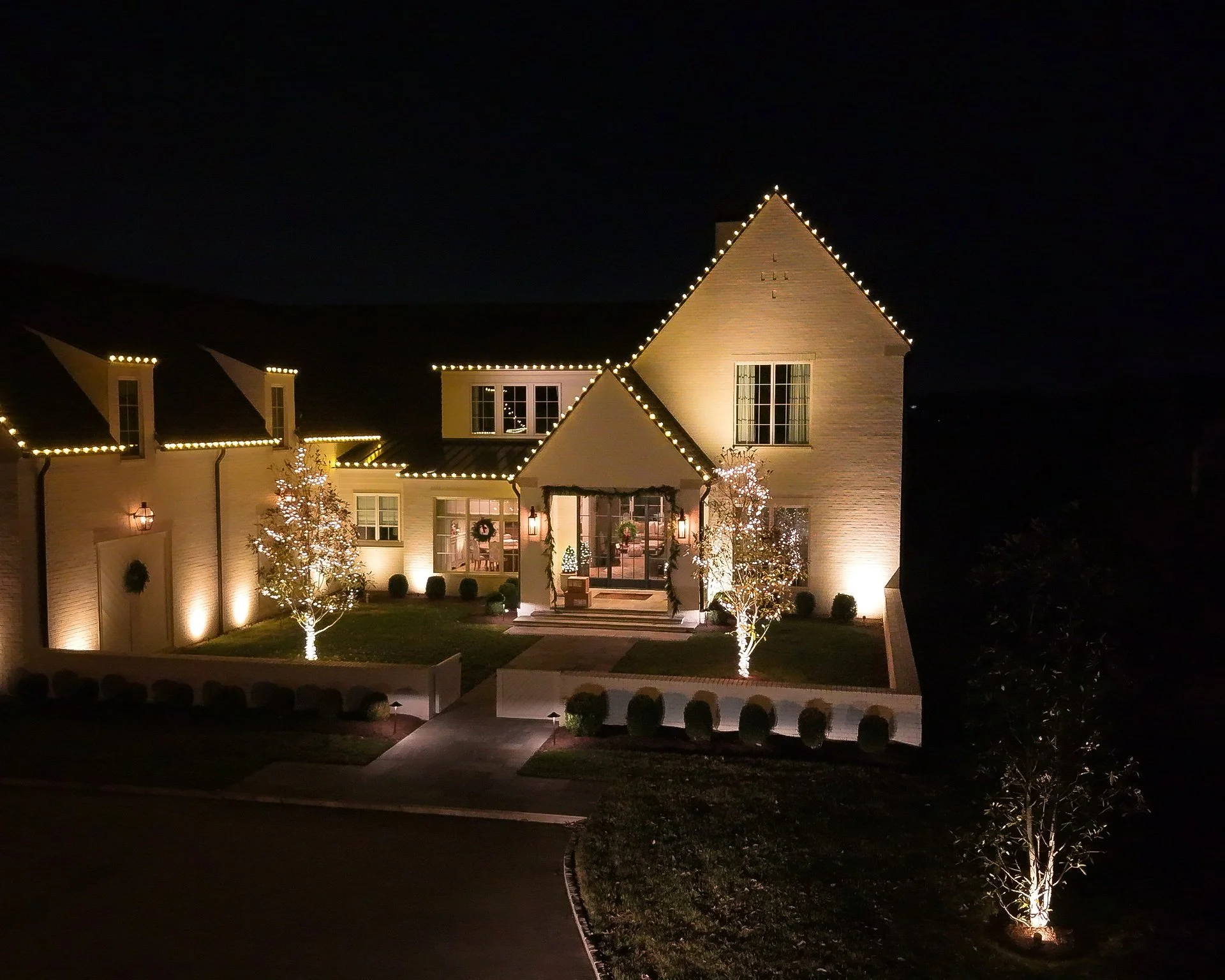 A well-lit house at night decorated with Christmas lights and wreaths, with some trees lit with string lights in the front yard.