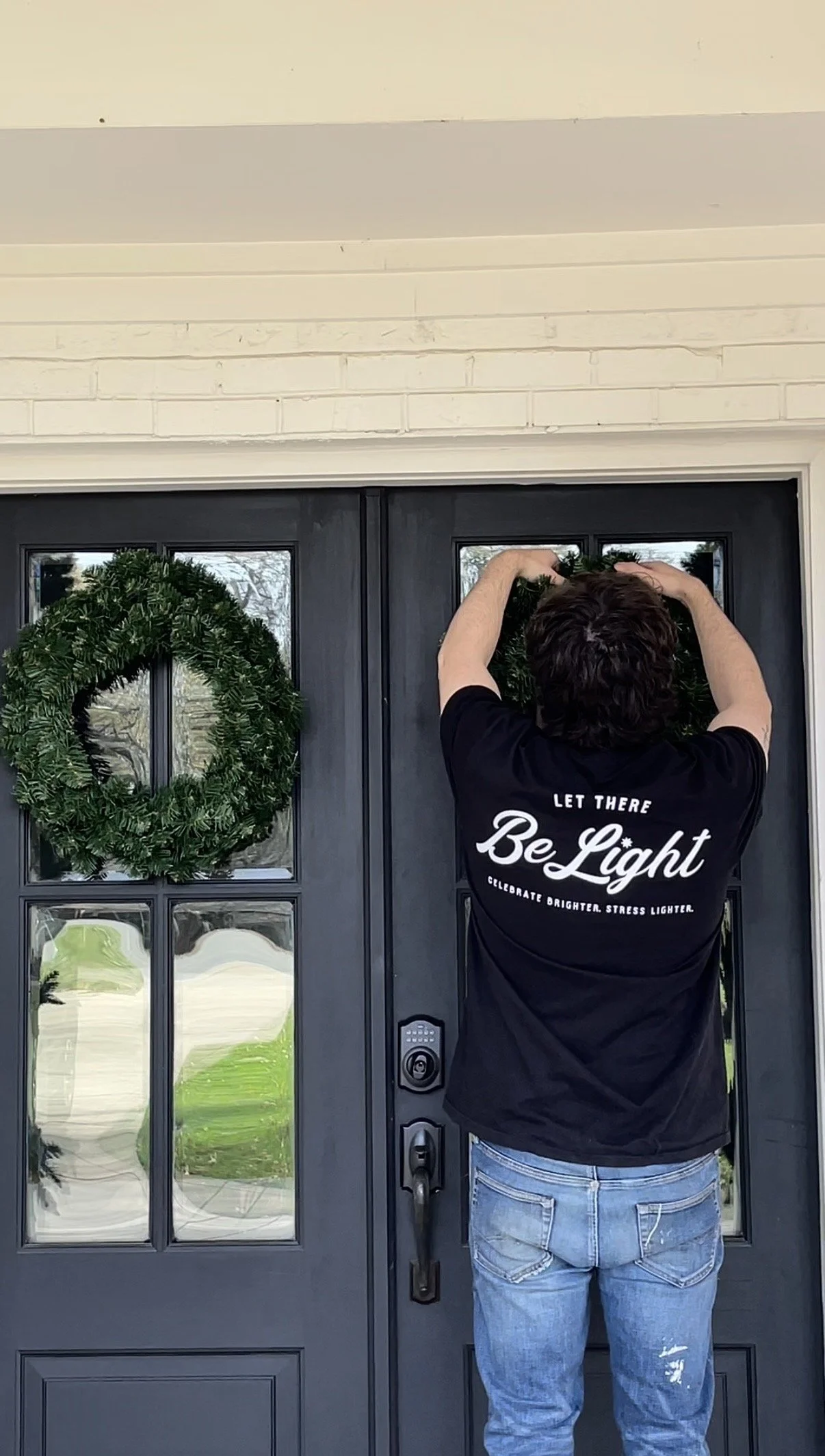 A person hanging a holiday wreath on a black front door.