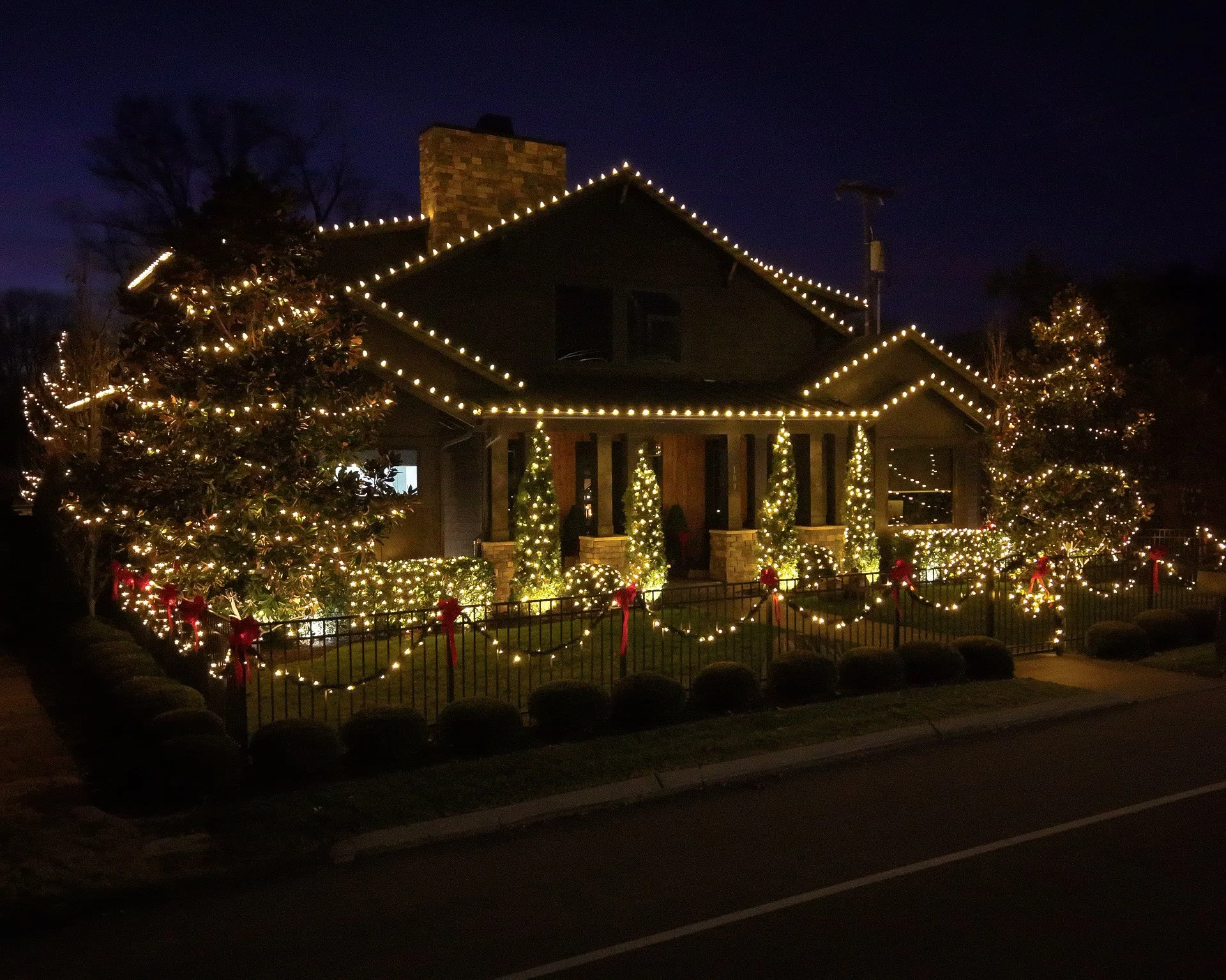 A house decorated for Christmas with lights outlining the roof, trees, and porch, surrounded by a fence decorated with bows and lights, at night.