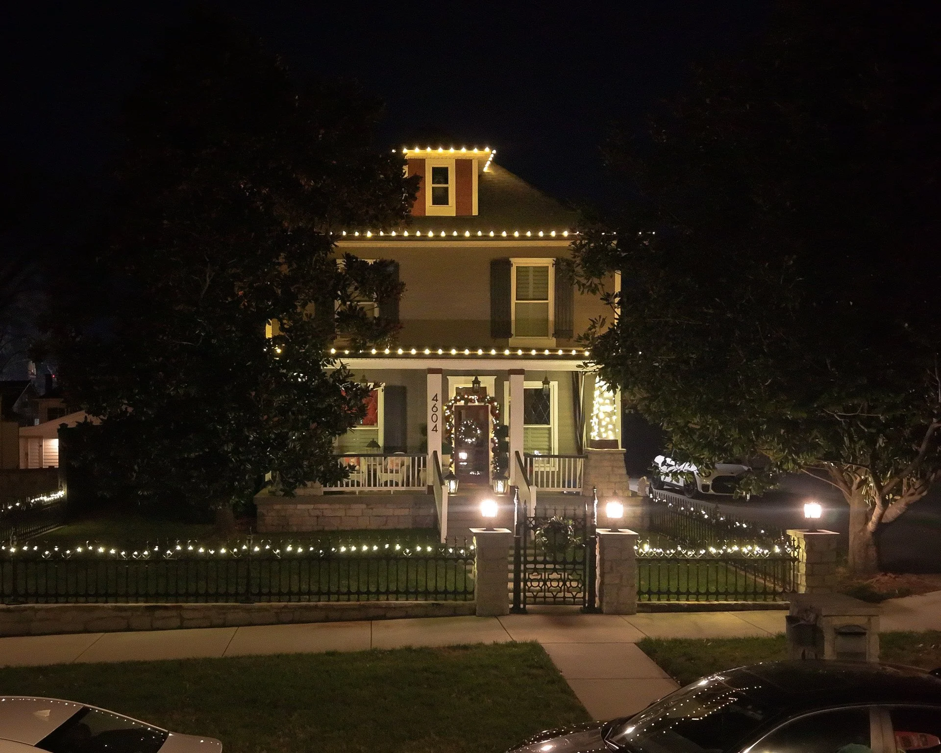 House decorated with Christmas lights at night, with two large trees in the front yard and parked cars nearby.